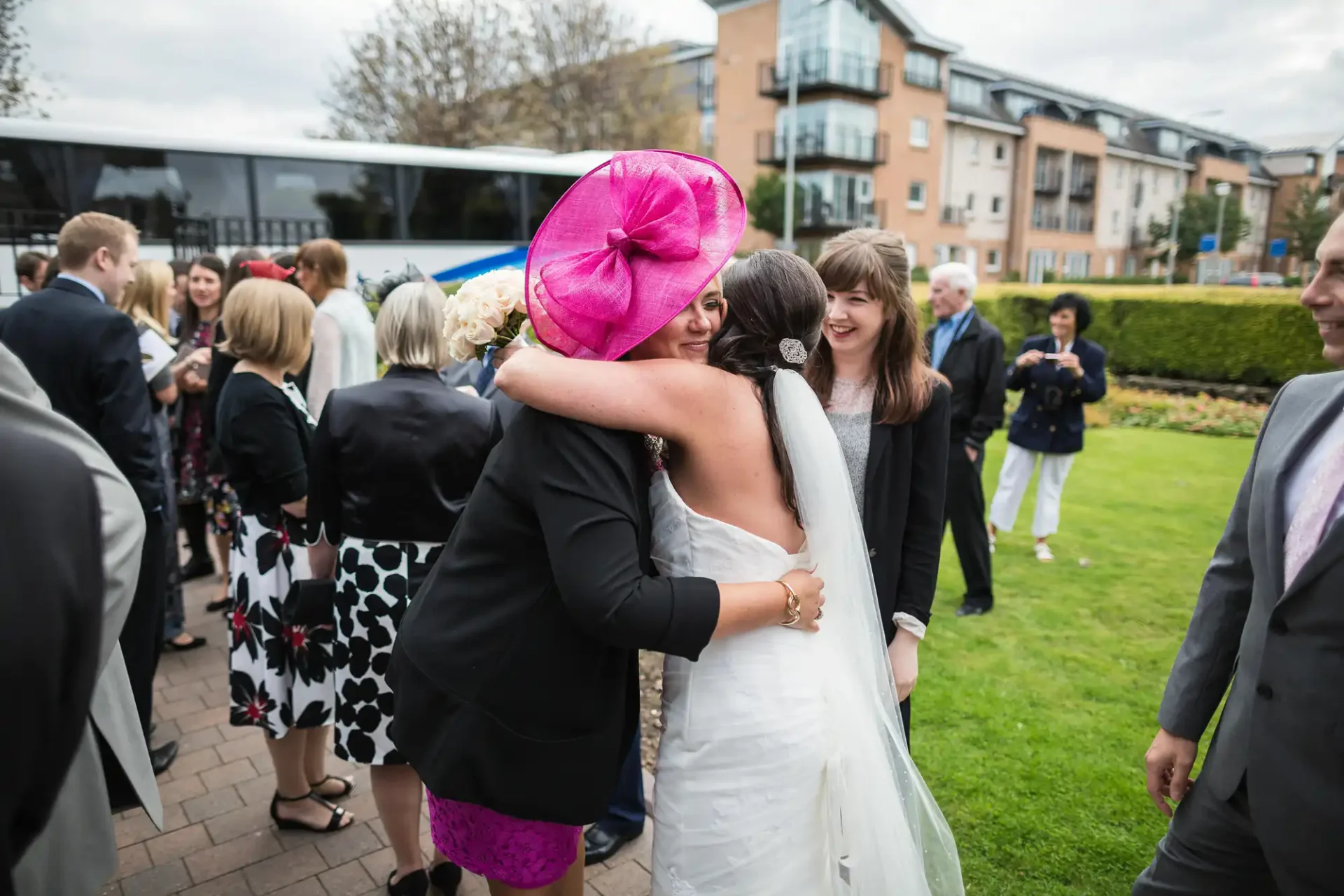 A bride in a white dress embracing a woman in a bright pink hat at a wedding, with guests nearby.