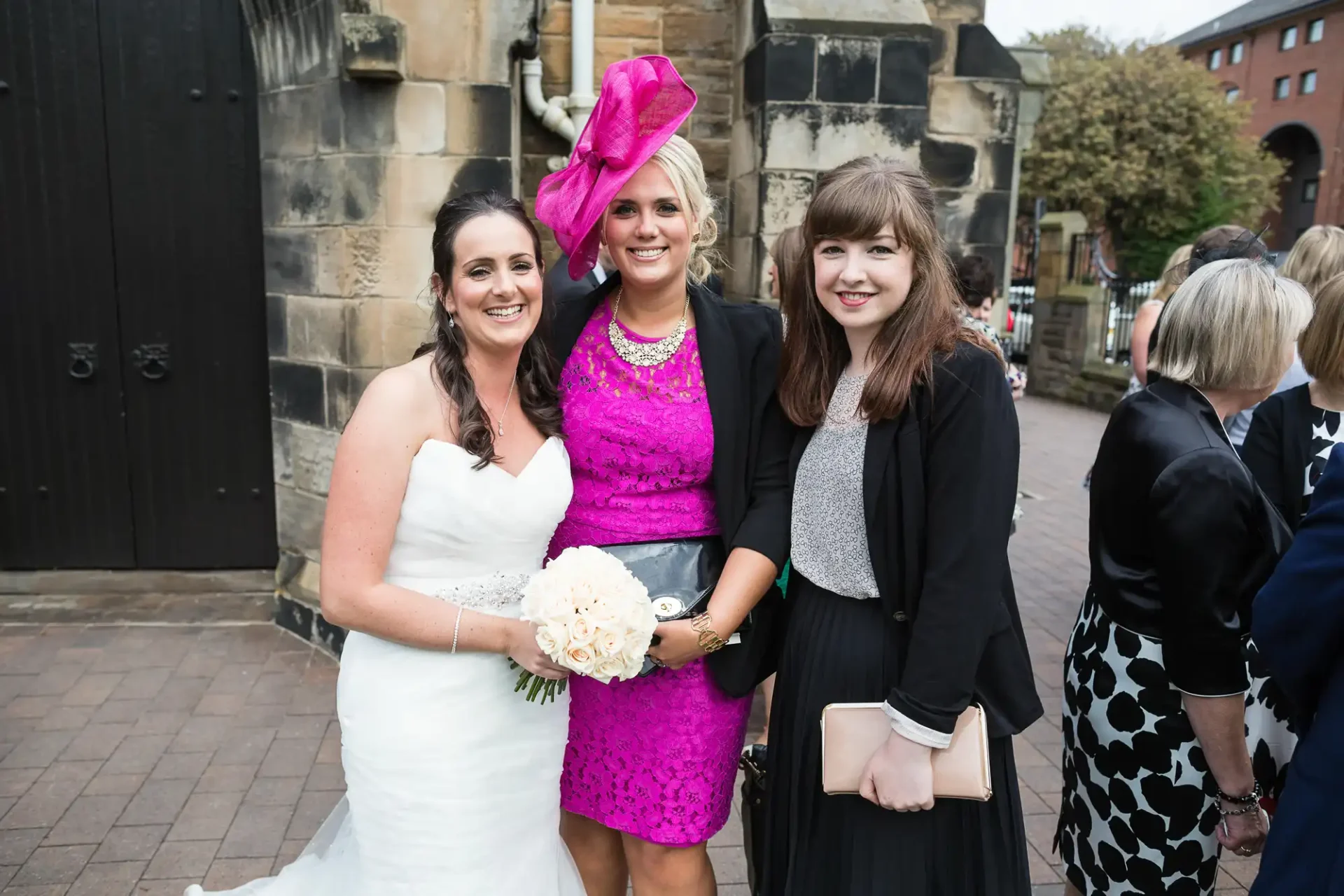 Bride in white dress with two women, one in a pink dress and large hat, and the other in a black cardigan, all smiling outdoors.