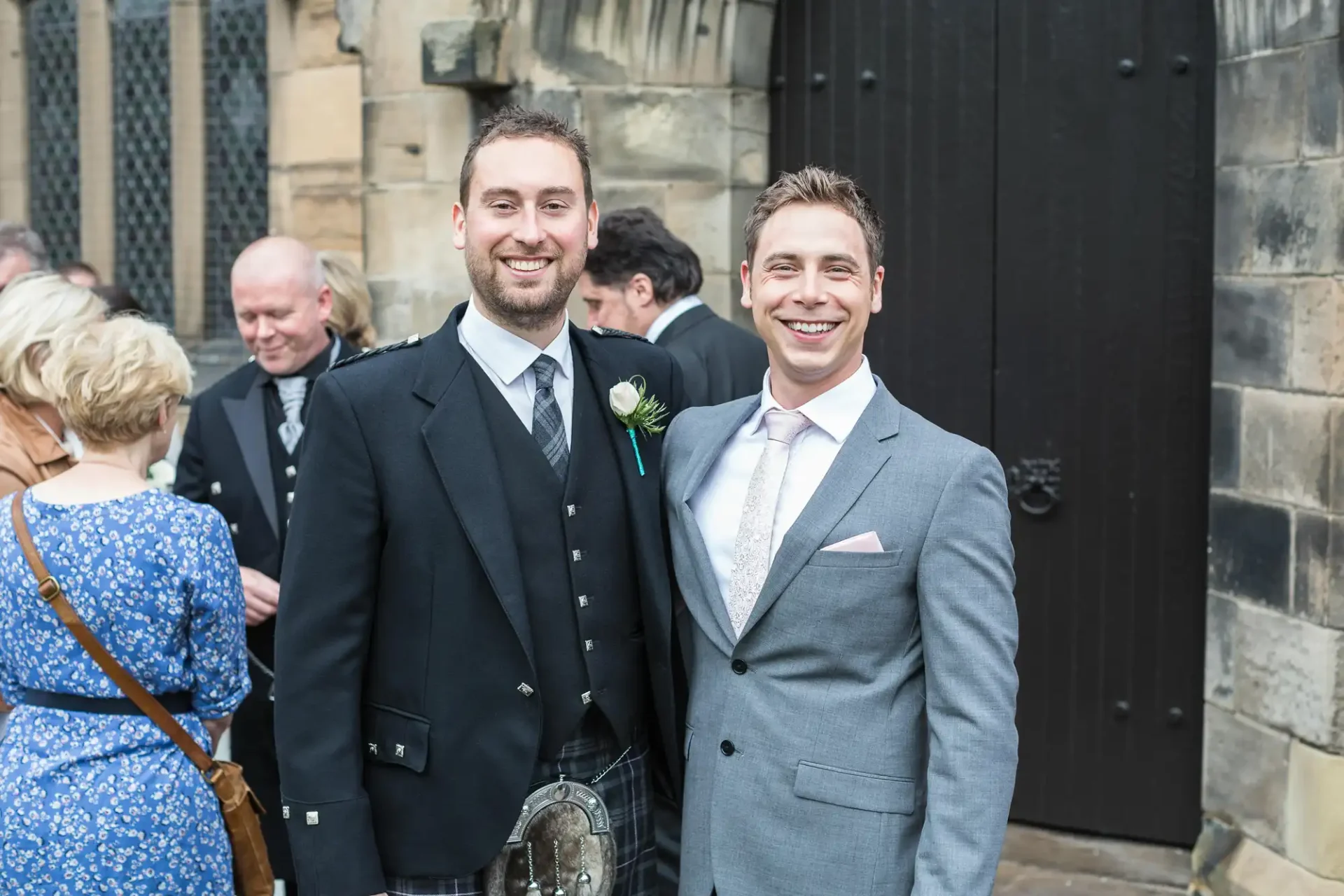 Two smiling men, one in a kilt and the other in a suit, standing together at a social gathering outside a building with a black door.