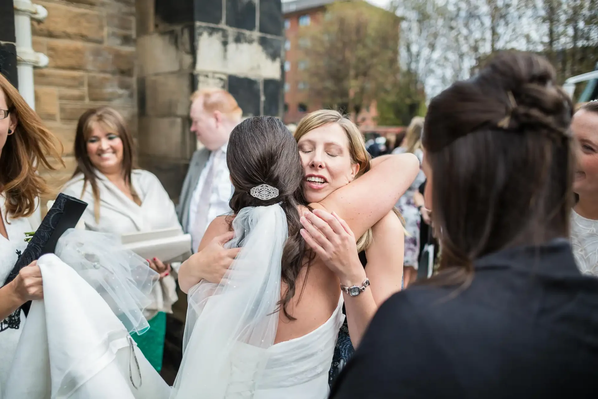 A bride and a woman warmly embracing at a wedding, with guests looking on and smiling.