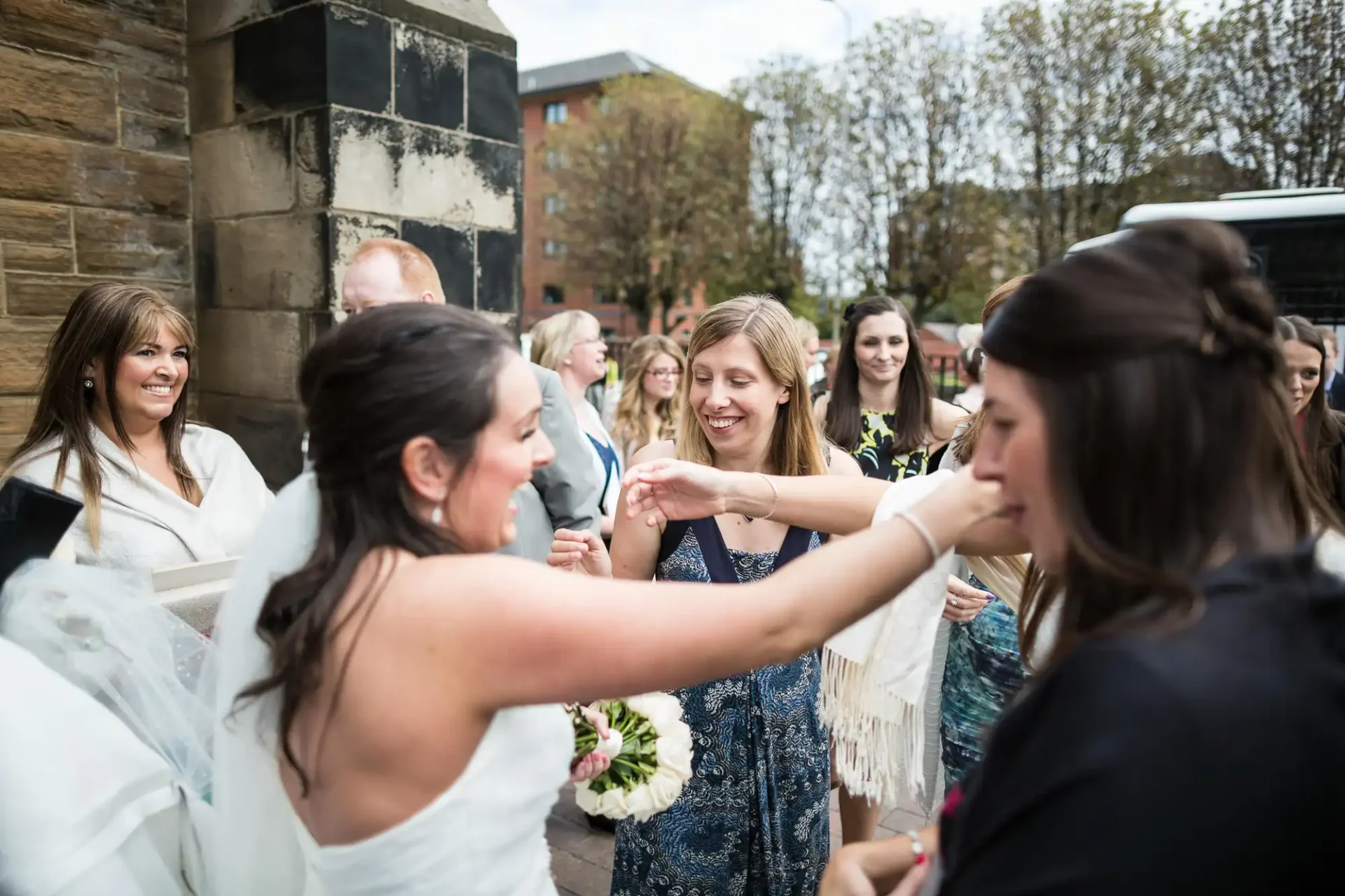 A bride in a white dress excitedly greets guests outside a building, with several smiling people surrounding her.