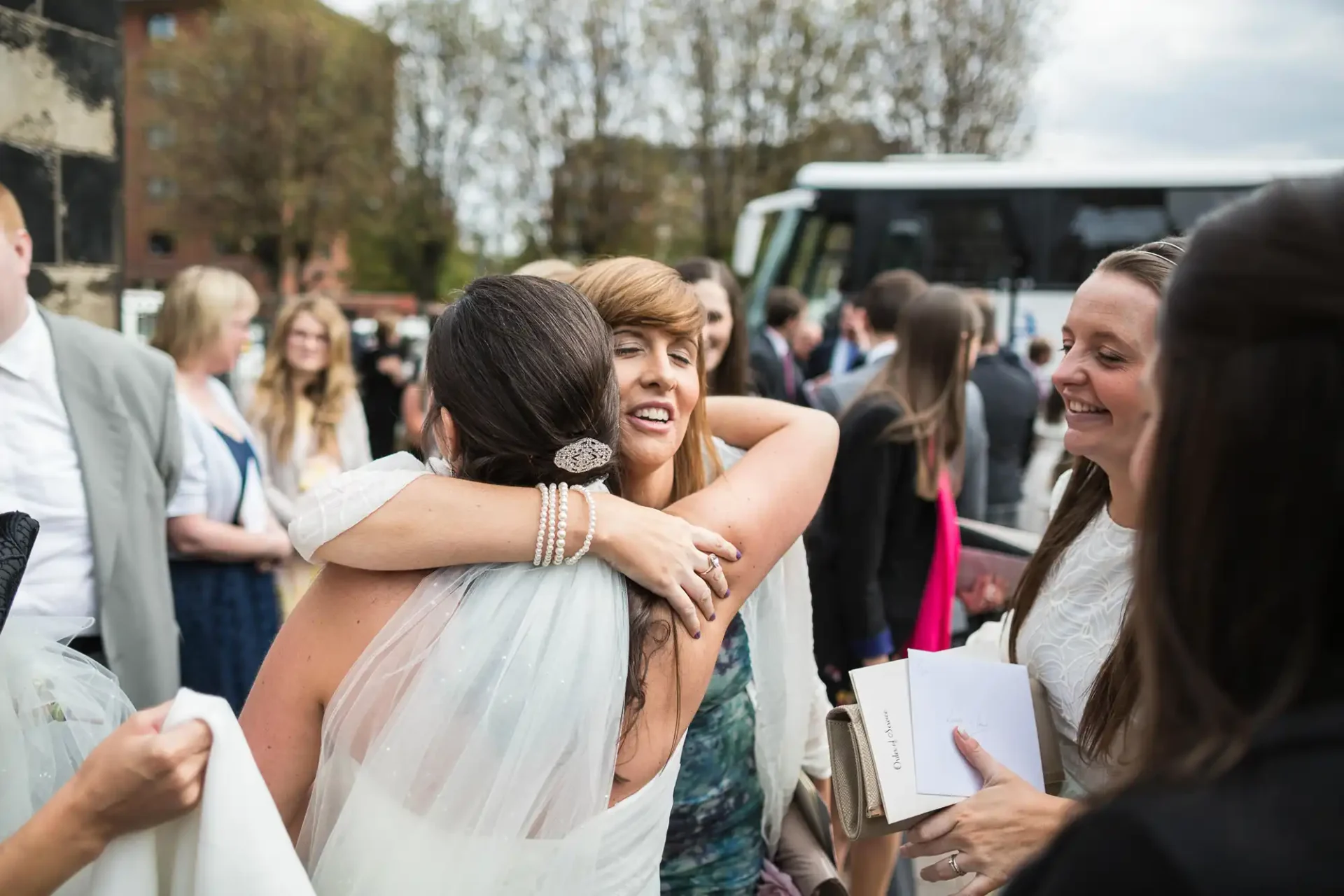 A bride hugs a guest at her wedding while other guests look on and smile in a lively outdoor gathering.