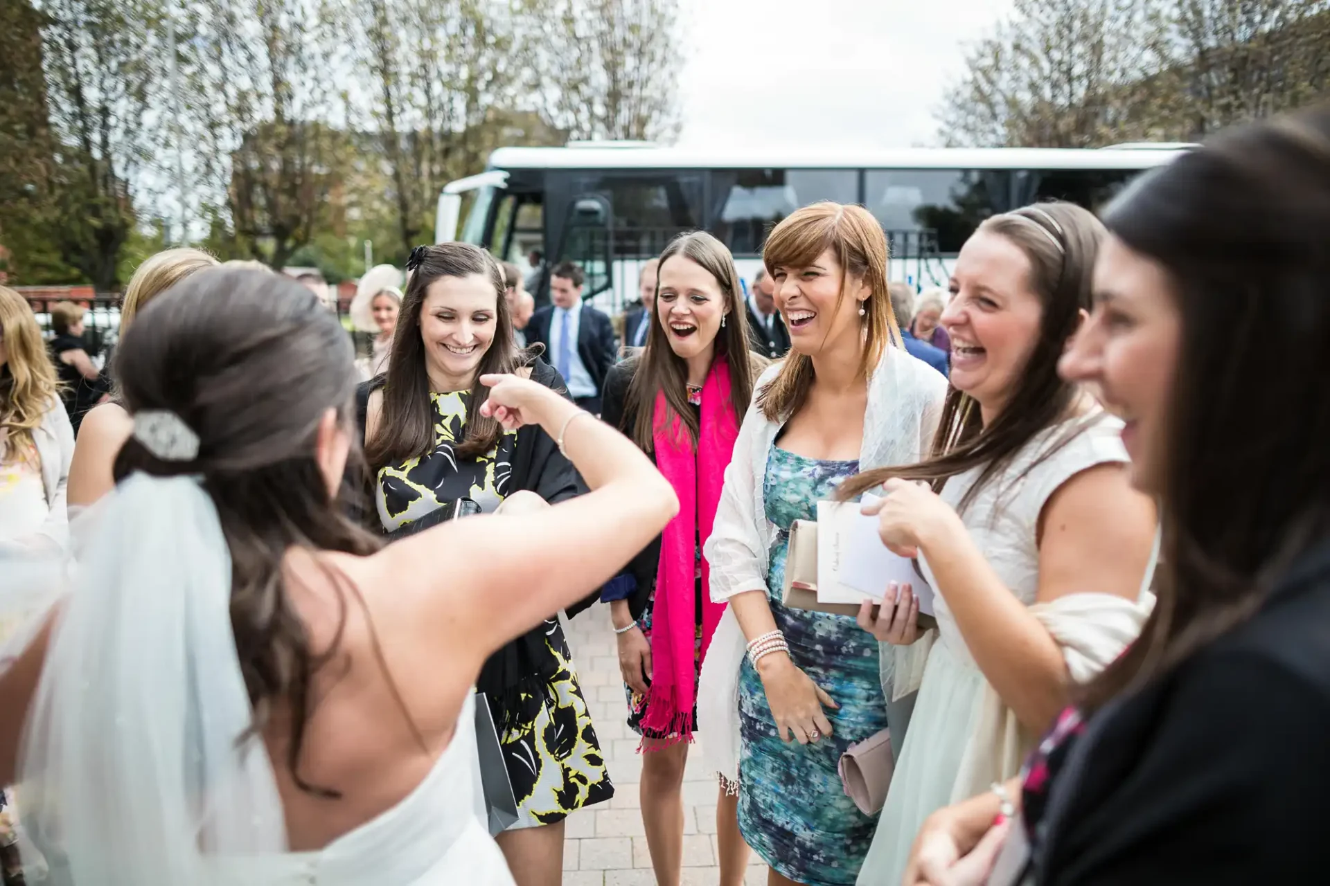 A bride joyfully greeting a group of women in colorful dresses at an outdoor event, with a bus parked in the background.