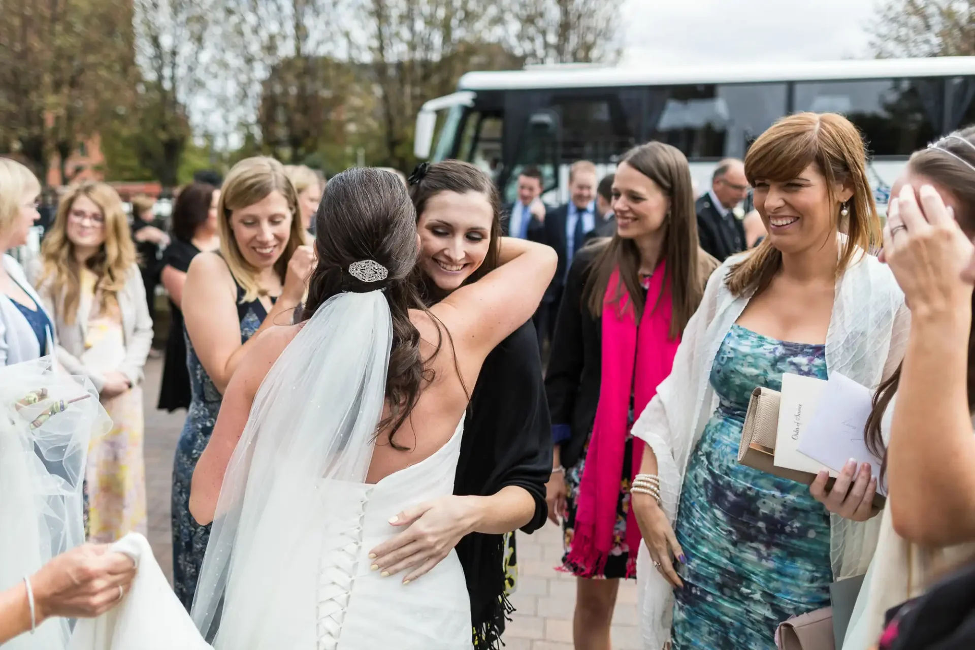 A bride embraces a guest at her wedding, surrounded by smiling female friends and family outdoors.