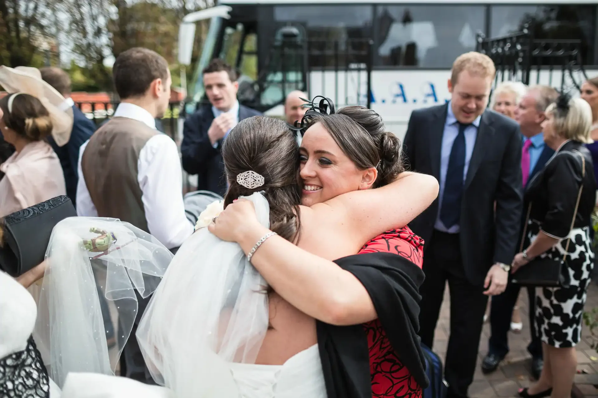 A bride in a white dress receives a warm embrace from a woman in a black and red outfit at a wedding gathering with guests around them.