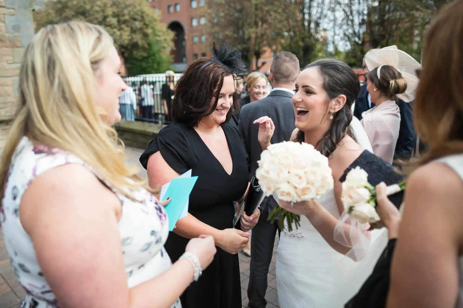 A bride holding a bouquet laughs joyfully with two women during a wedding celebration outdoors.