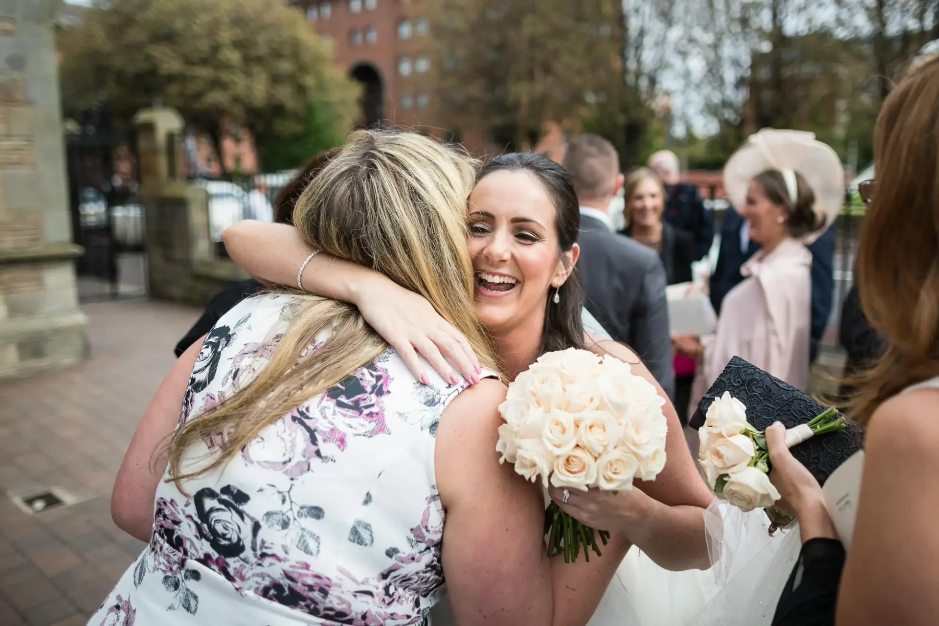 A bride with a bouquet of white roses hugging a woman in a floral dress at a wedding ceremony, with guests smiling in the background.