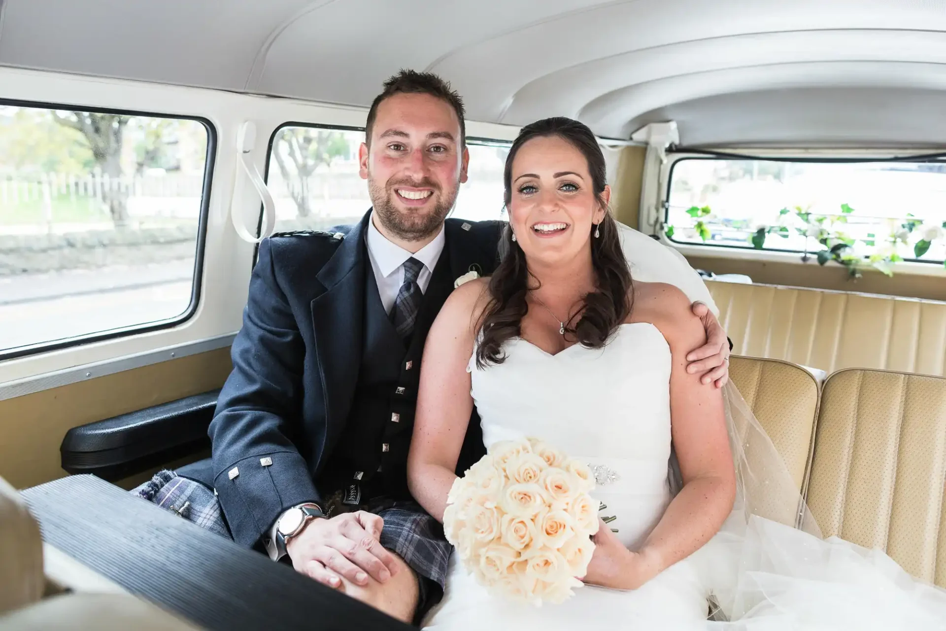 A newlywed couple smiling inside a vintage car, the groom in a kilt and the bride holding a bouquet of roses.