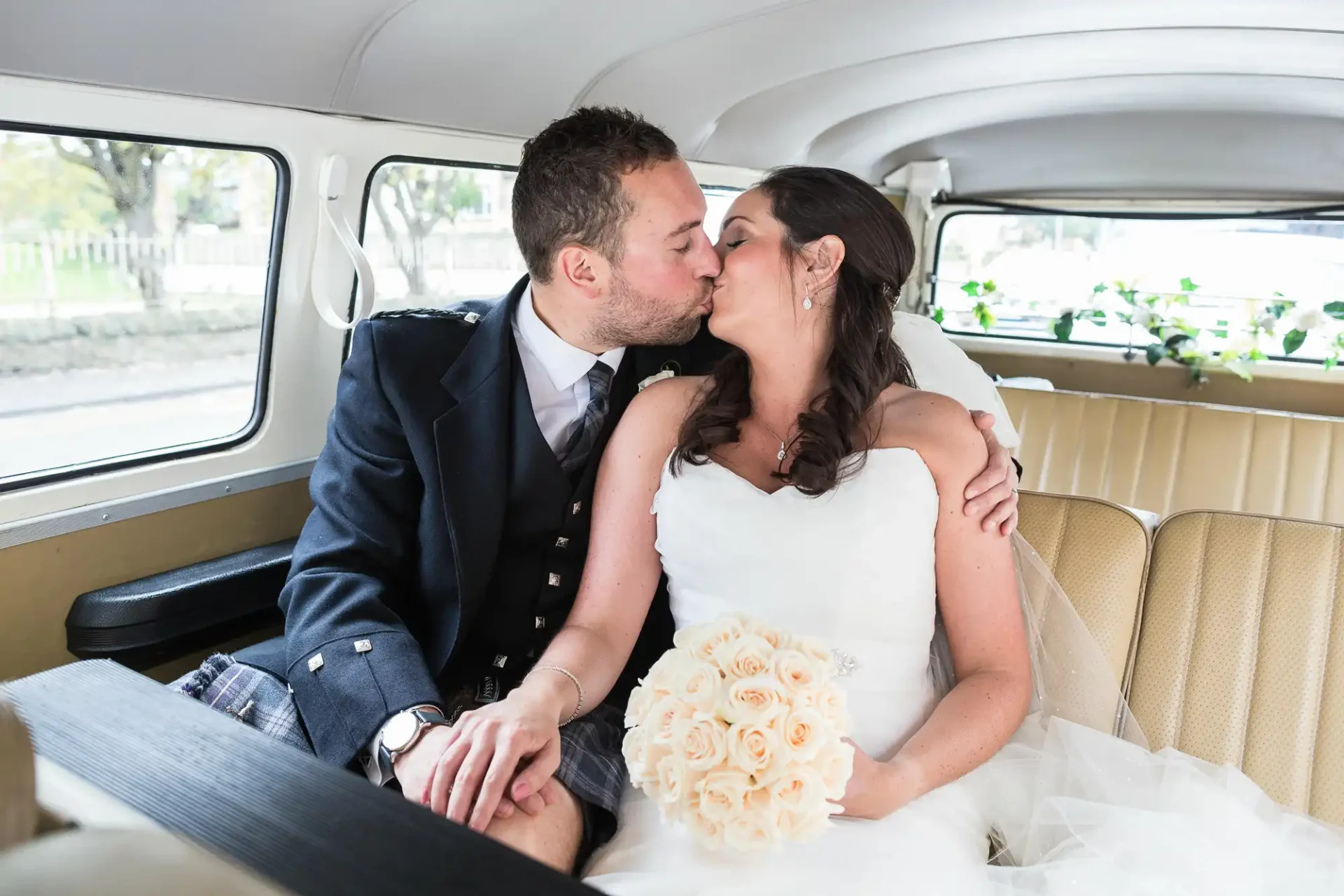 Bride and groom kissing inside a classic car, with the bride holding a bouquet of cream roses.