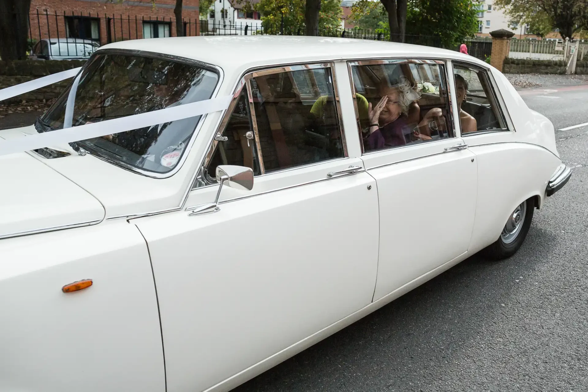White wedding limousine with ribbons, carrying passengers visible through the windows, parked on a tree-lined street.