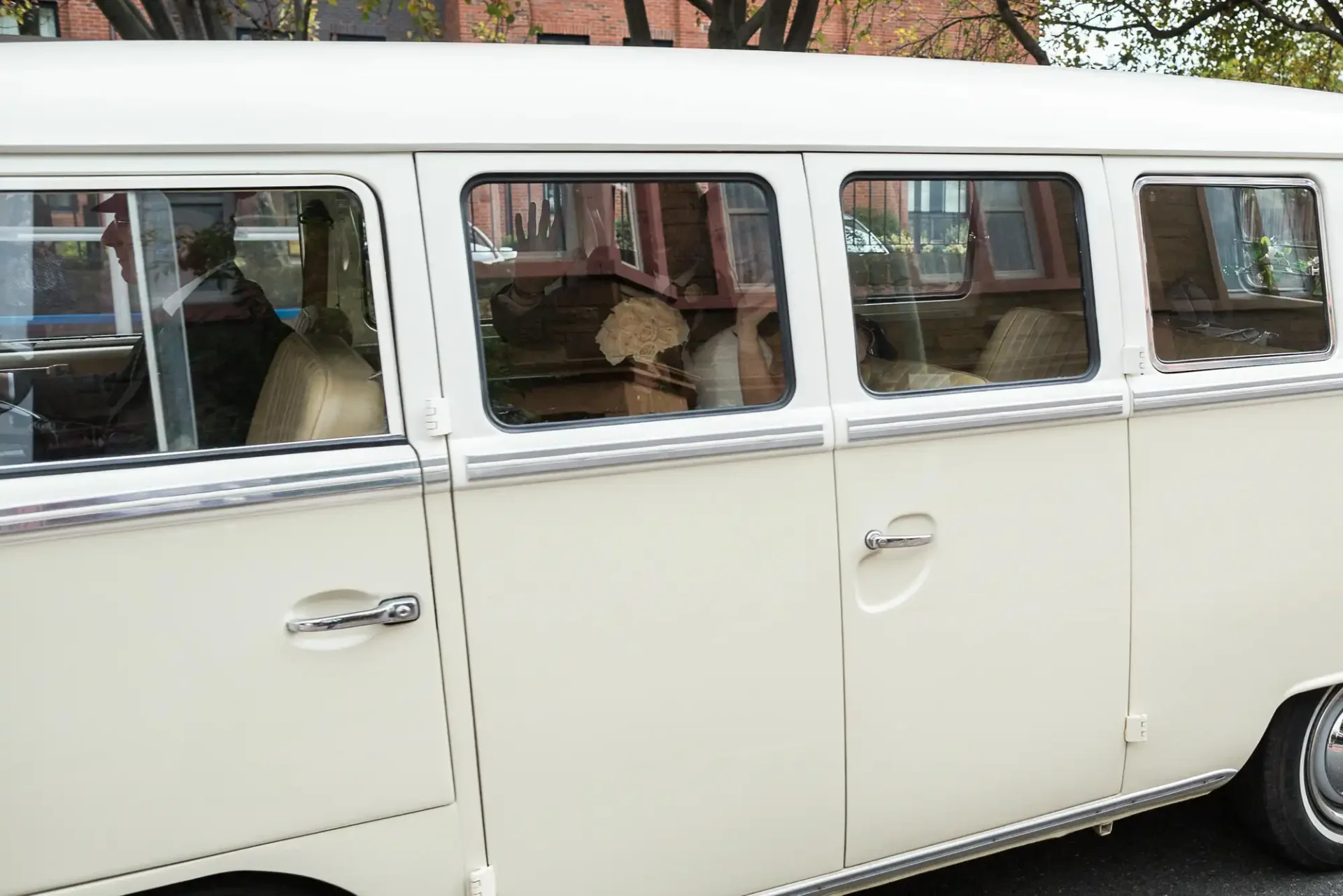 Side view of a cream-colored vintage volkswagen bus with multiple windows, parked on a city street.