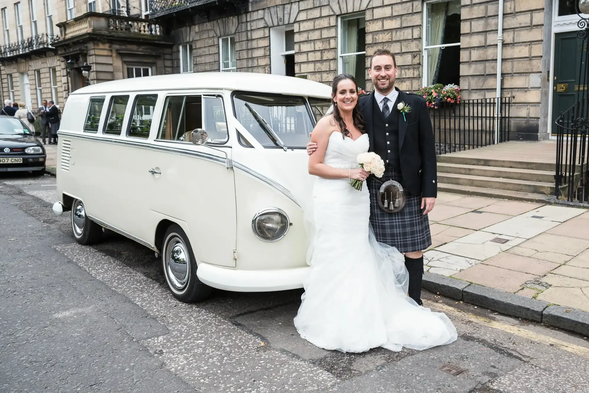 Bride in a white dress and groom in a kilt standing next to a classic white volkswagen van on a city street.