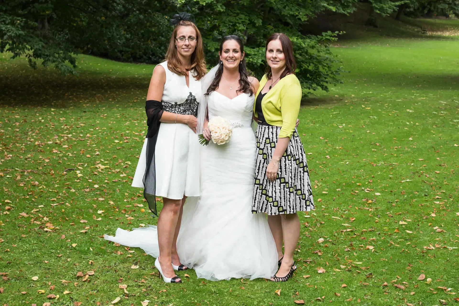 Three women smiling in a park, one in a white bridal gown flanked by two in stylish dresses, with trees and grass in the background.