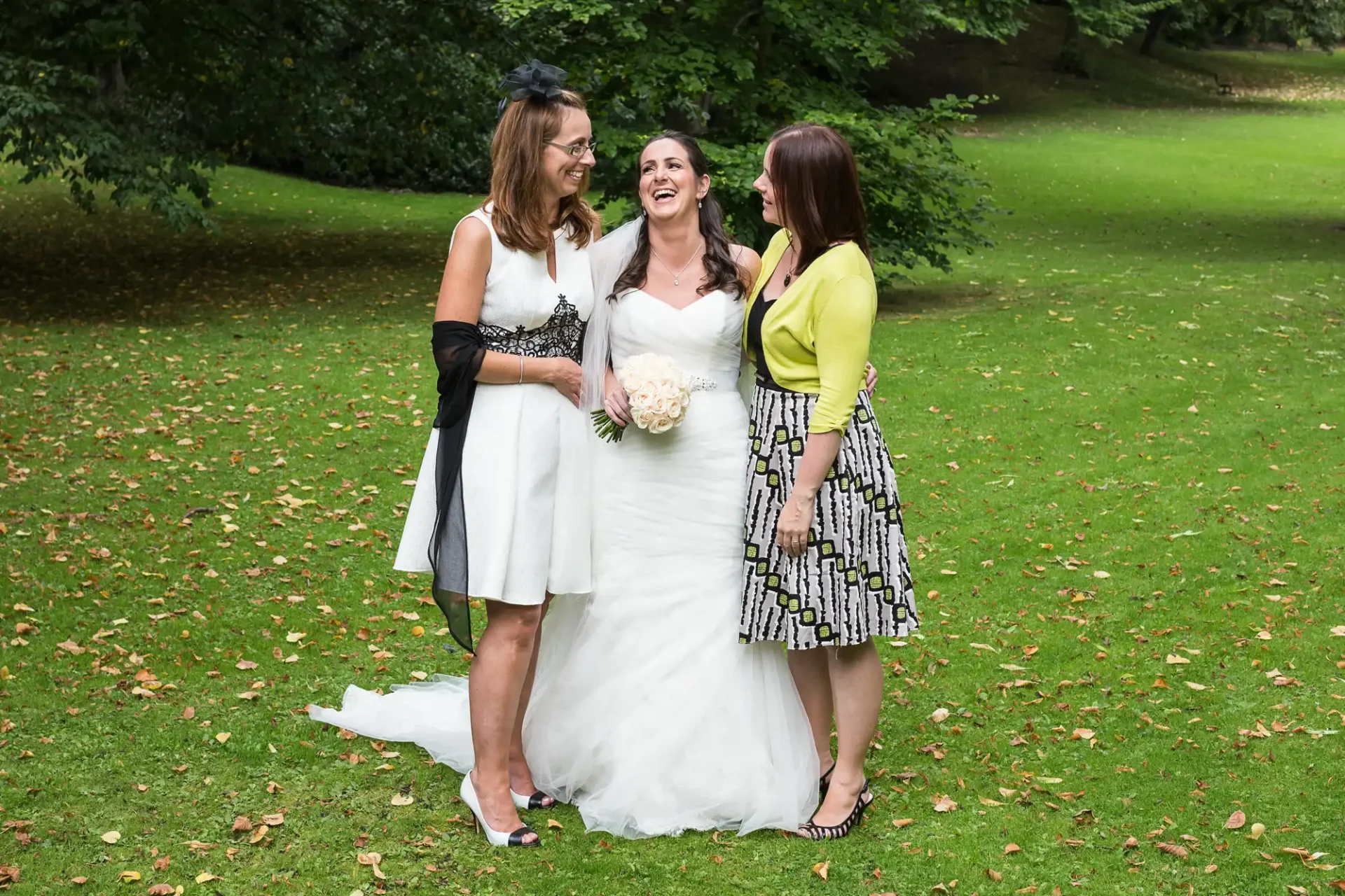 Three women, one in a white wedding dress, laugh together in a lush park. the other two wear stylish dresses, one black and white, the other yellow.