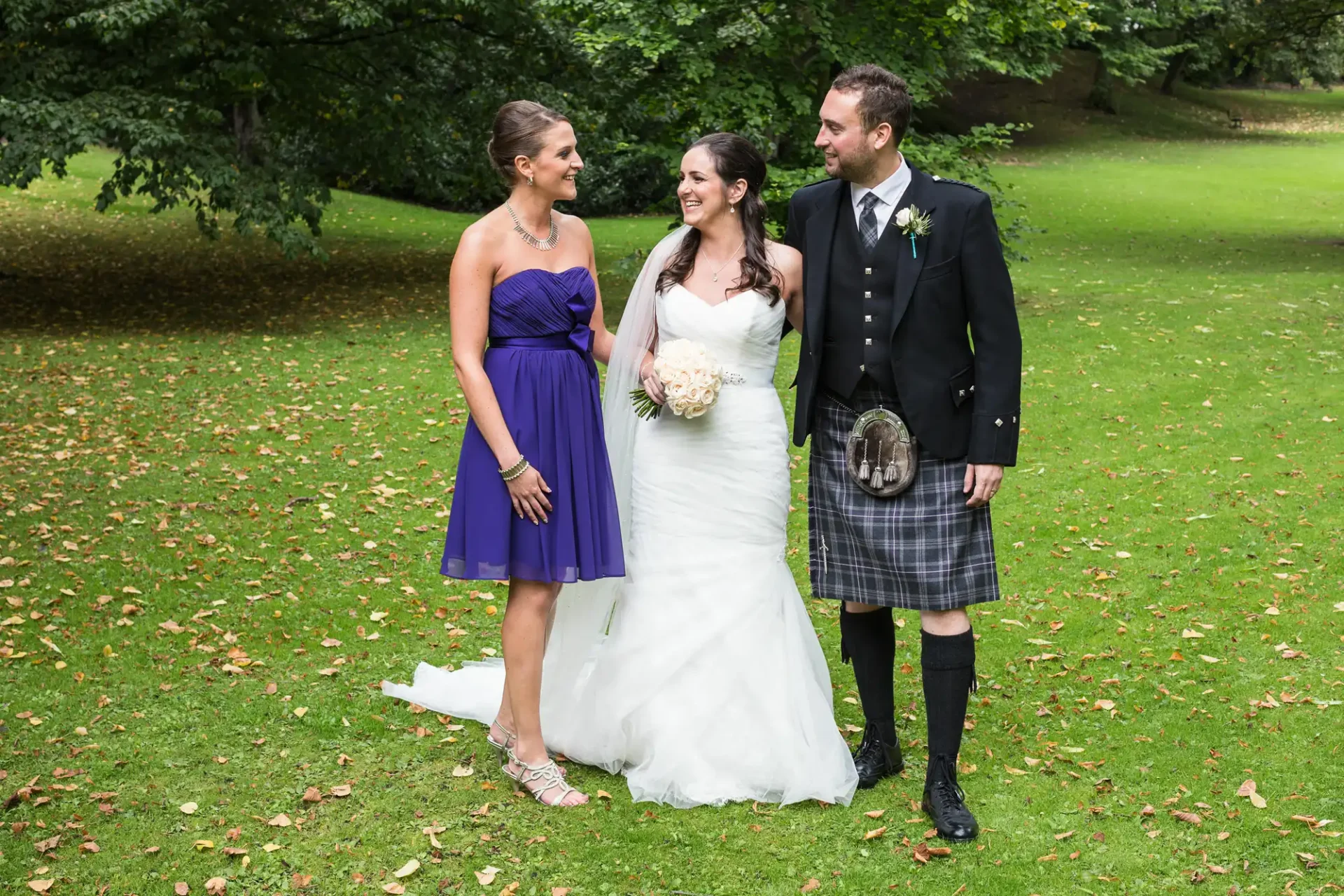 A bride in a white gown and a groom in a kilt stand with a bridesmaid in a purple dress, all smiling in a park with green trees.