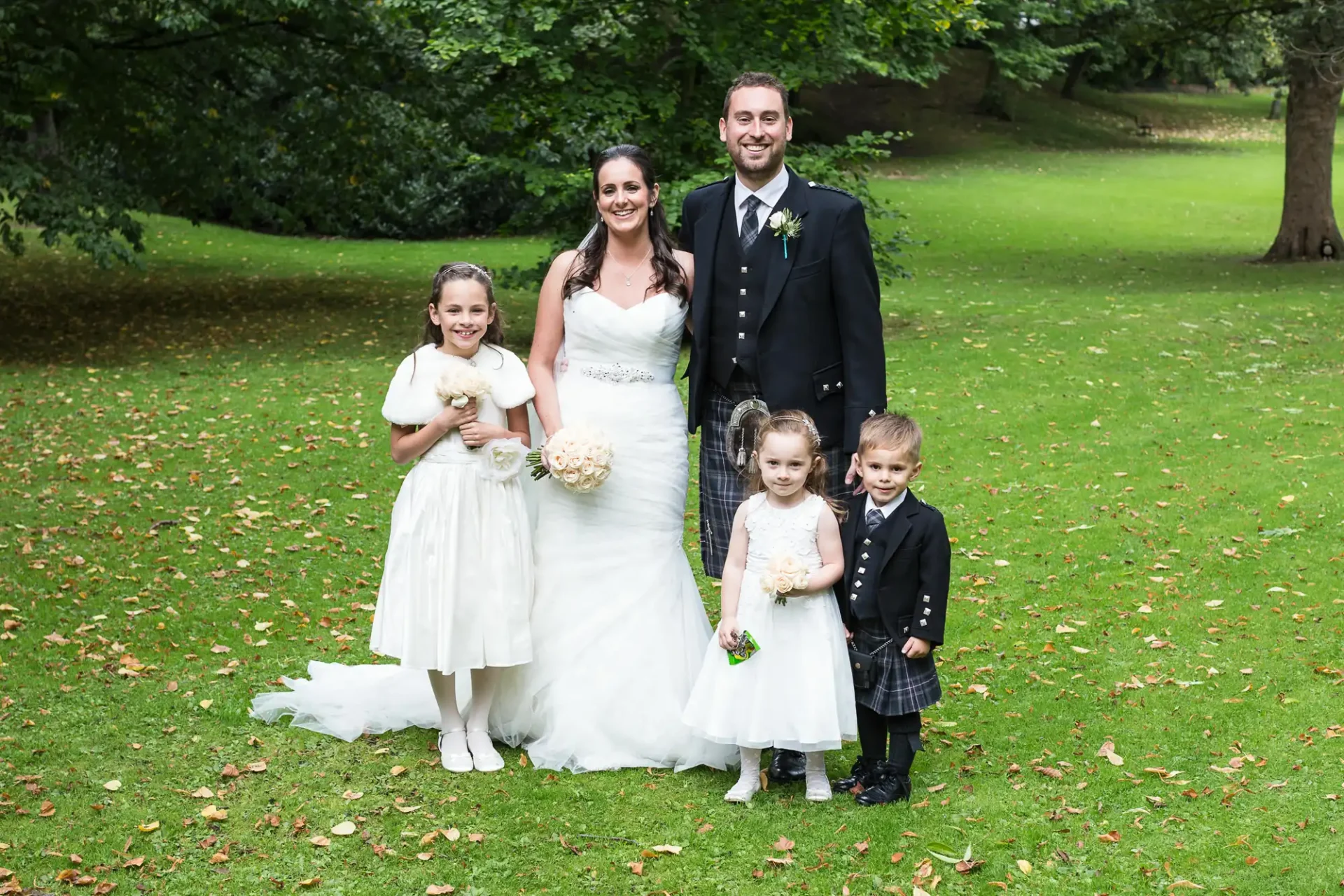 A bride and groom posing with three children in a park, the girls in dresses and the boy in a kilt, all smiling.