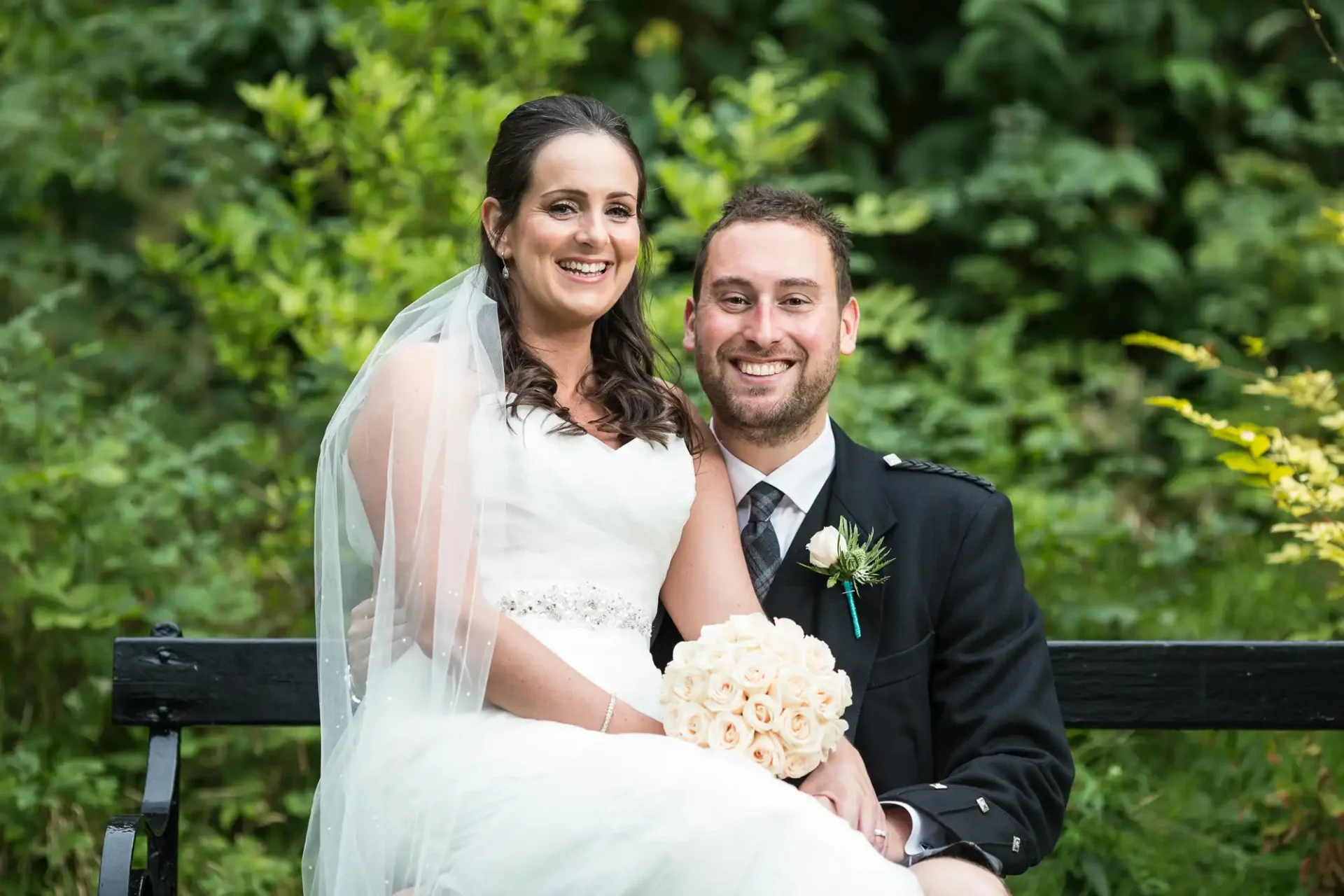 A smiling bride and groom seated on a park bench, the bride holding a bouquet of cream roses.