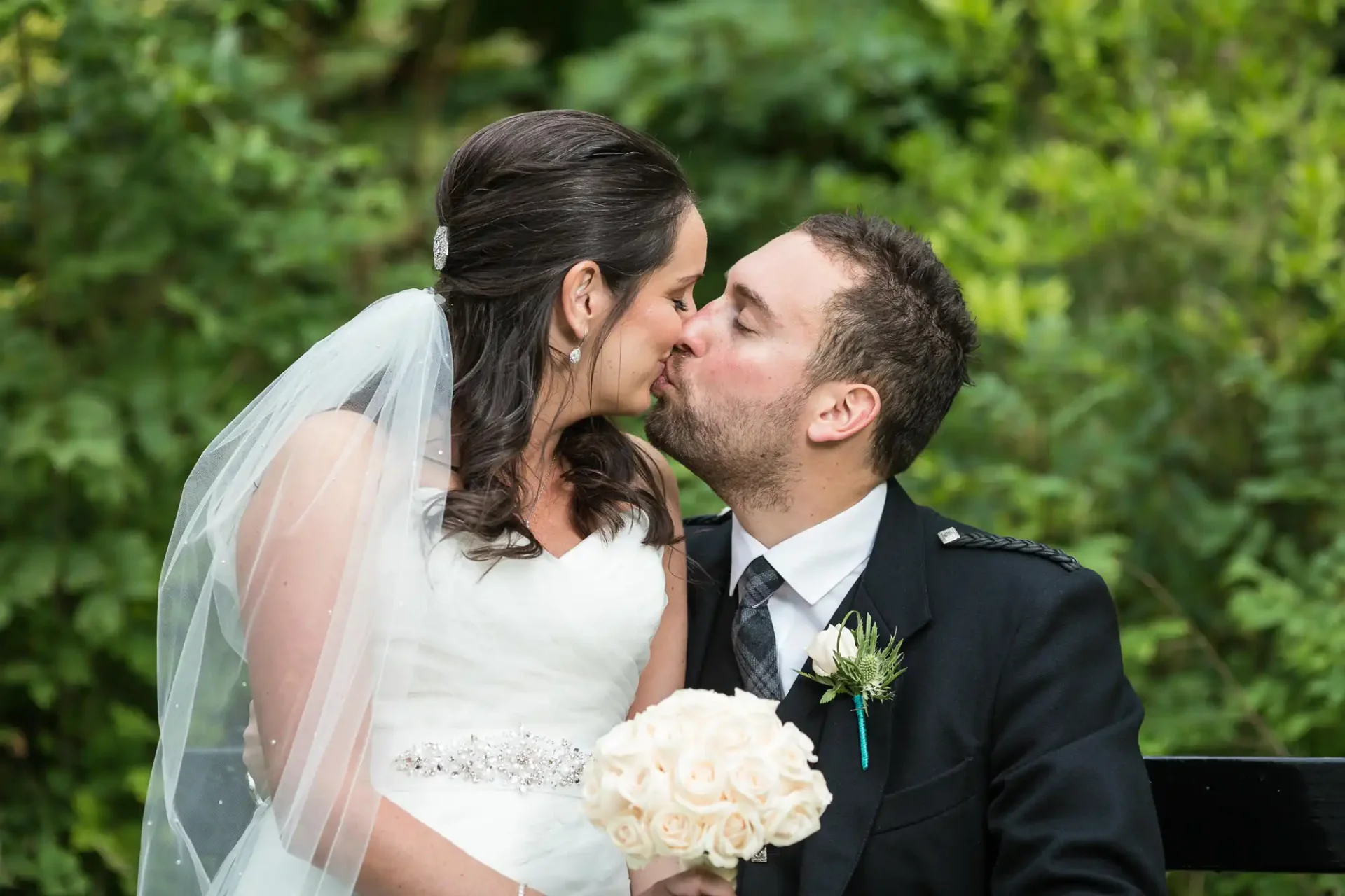 A bride and groom in wedding attire kissing in a lush green park, with the bride holding a bouquet of white roses.