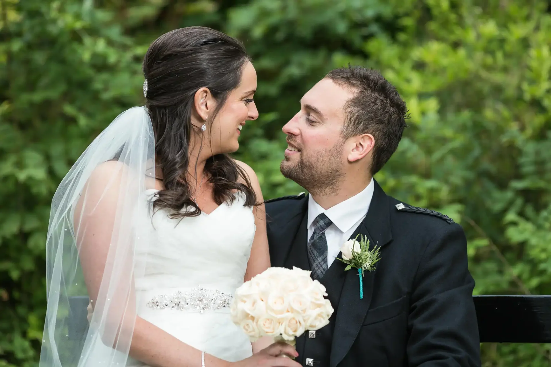 A newlywed couple happily looking at each other, the bride in a white gown holding a bouquet and the groom in a black suit, outdoors with lush greenery in the background.