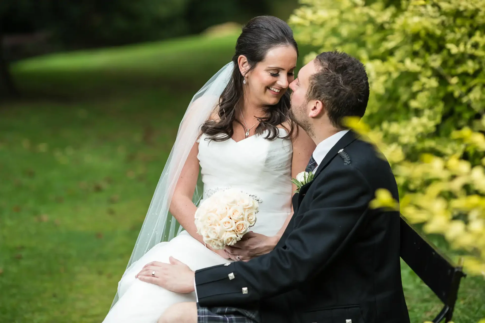 Bride and groom sitting on a park bench, smiling lovingly at each other, surrounded by greenery. the bride holds a bouquet of white roses.