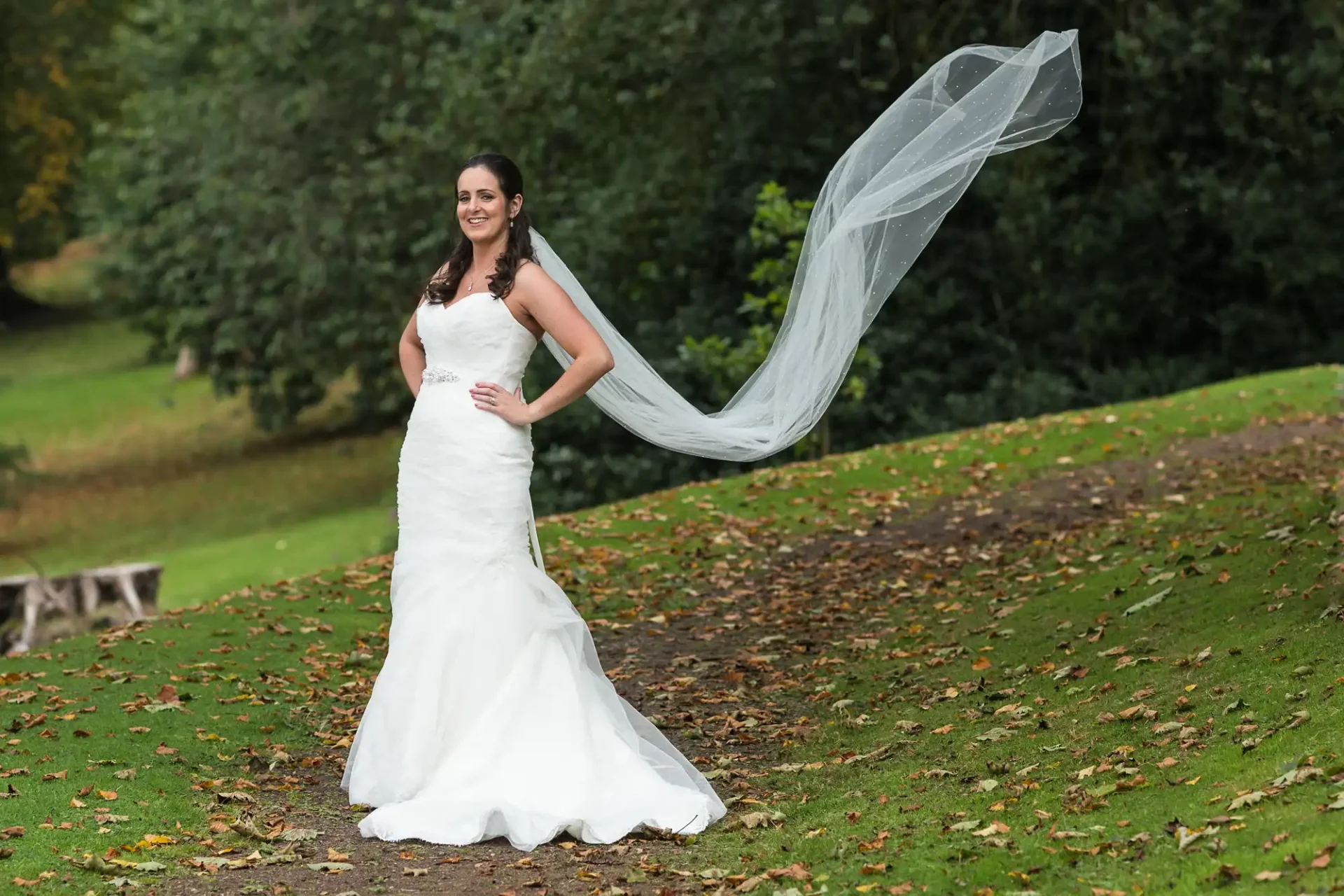 A bride in a strapless white gown smiles broadly, standing on a grassy slope with her long veil flowing elegantly in the breeze.