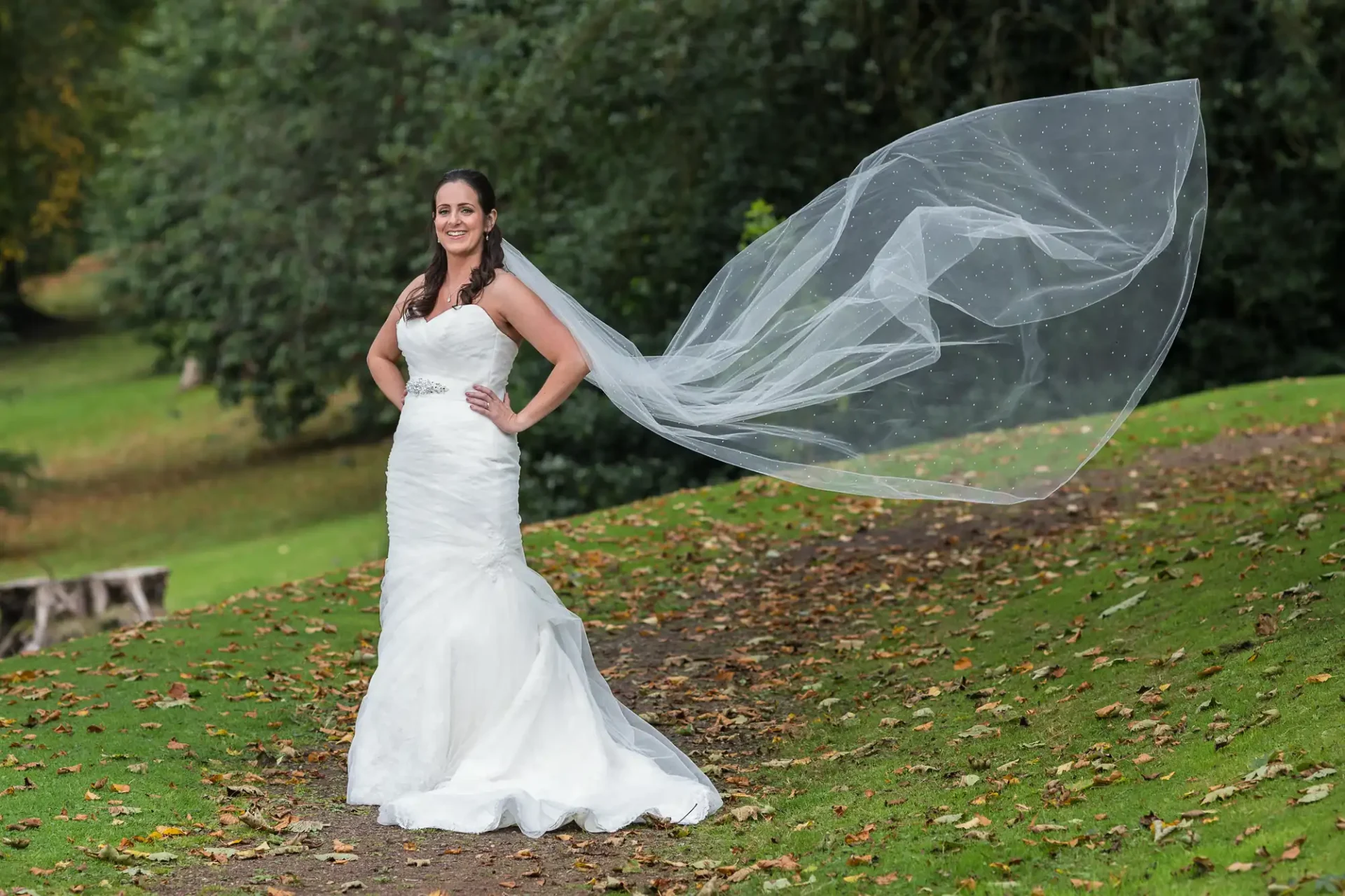 A bride in a strapless white gown stands on a grassy slope, smiling, with her long veil blowing in the wind.