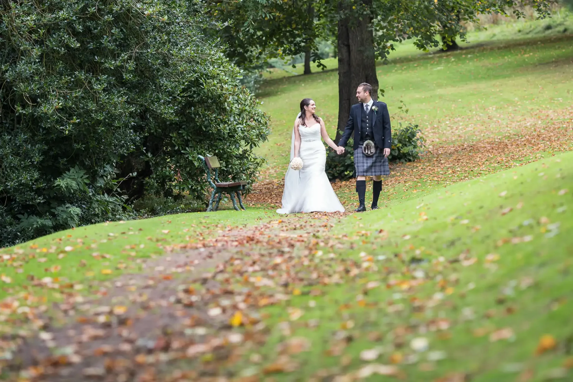 A bride and groom walking hand-in-hand in a lush park, with the groom wearing a kilt and the bride in a white gown.