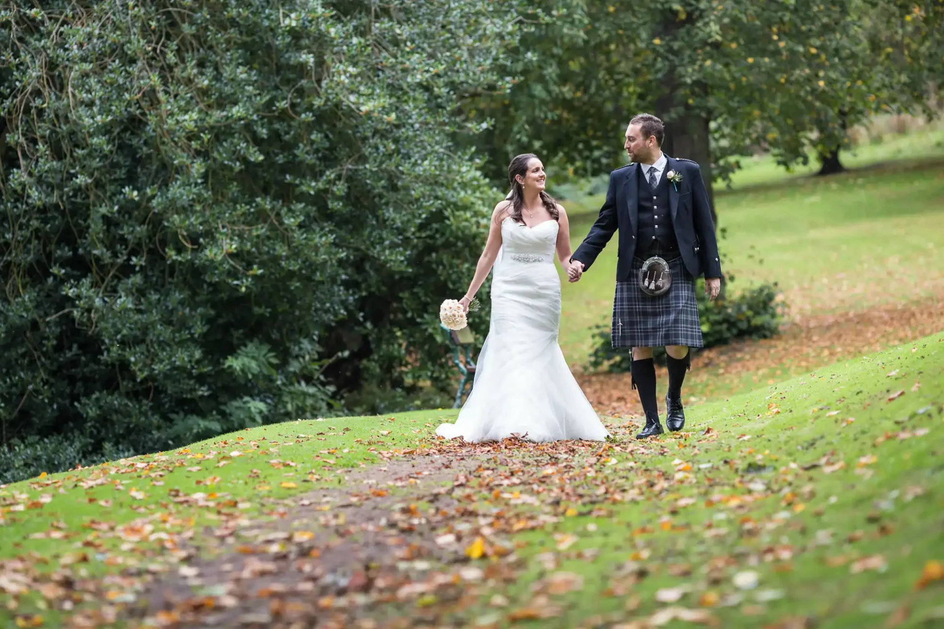 Bride in a white dress and groom in a kilt walking hand-in-hand down a leaf-strewn path in a park.