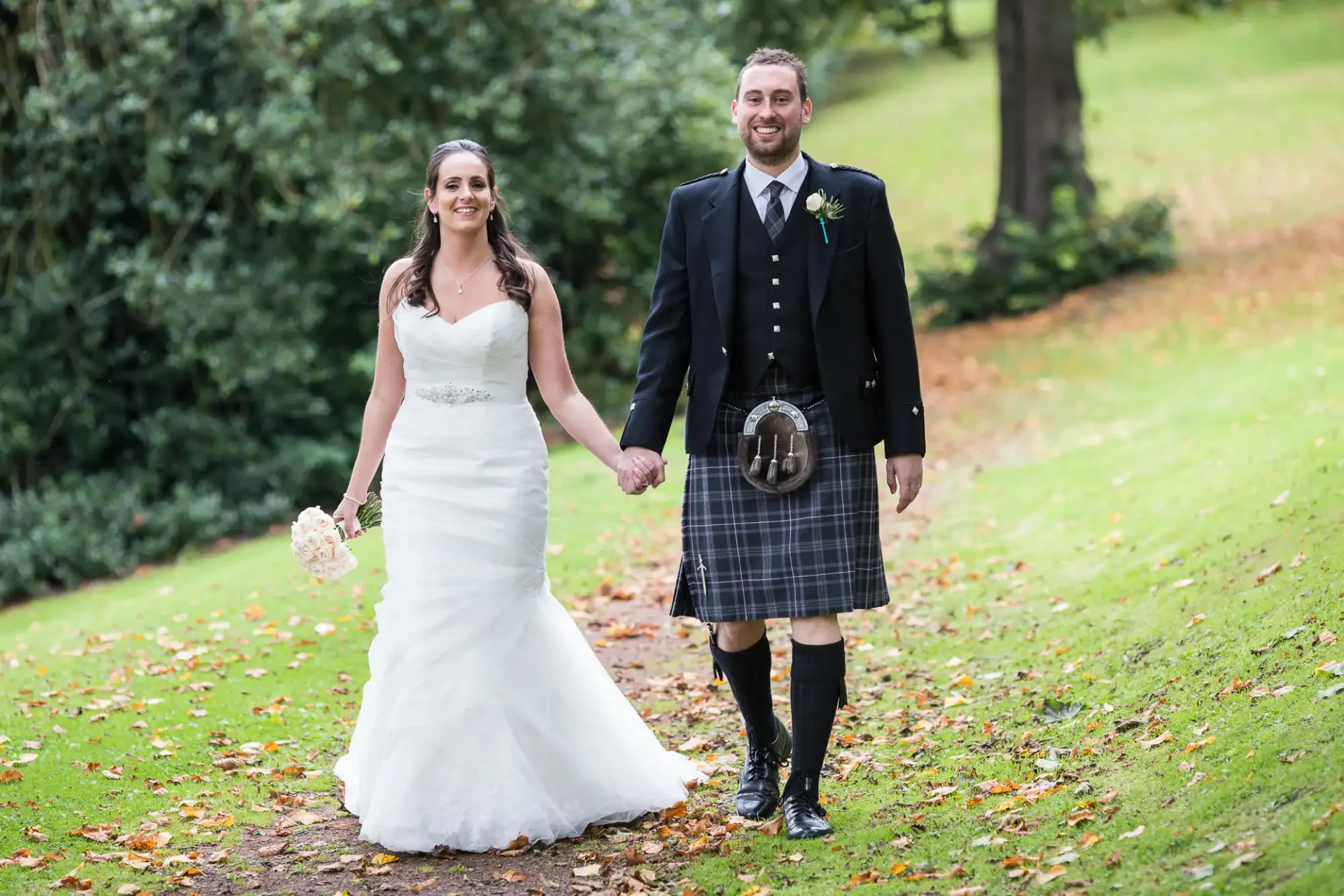 A bride in a white dress and a groom in a kilt holding hands and smiling while walking through a park with scattered fallen leaves.