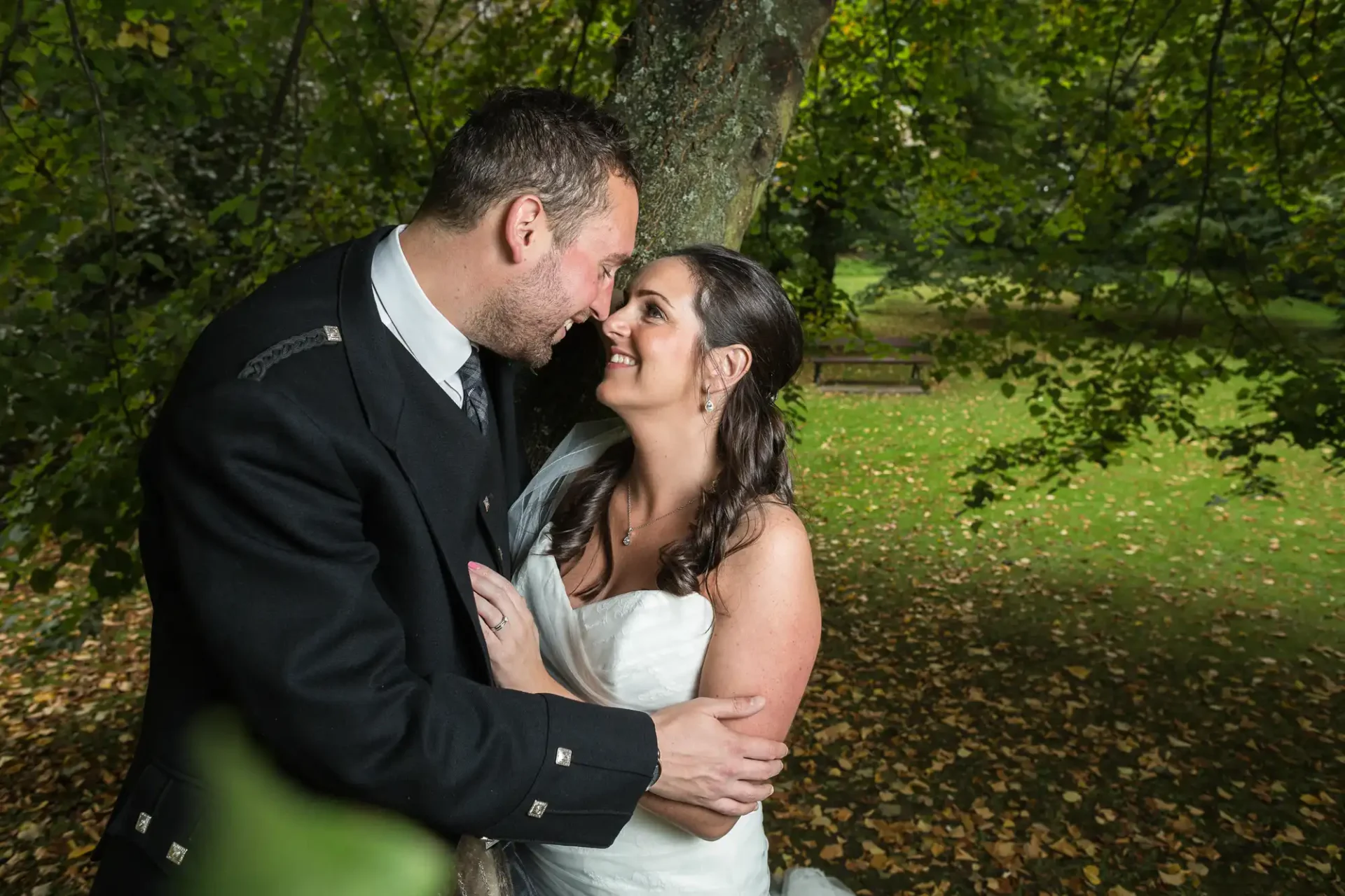 A bride and groom embrace affectionately in a lush, green park, with sunlight filtering through the trees.