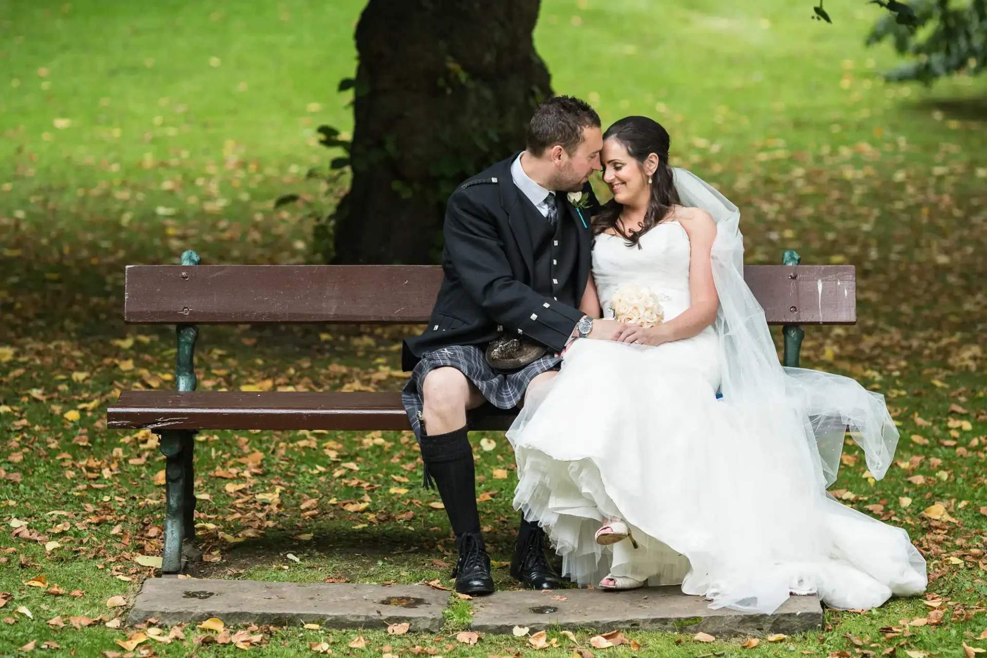 A bride in a white dress and a groom in a kilt sitting on a park bench, affectionately touching foreheads under a tree.