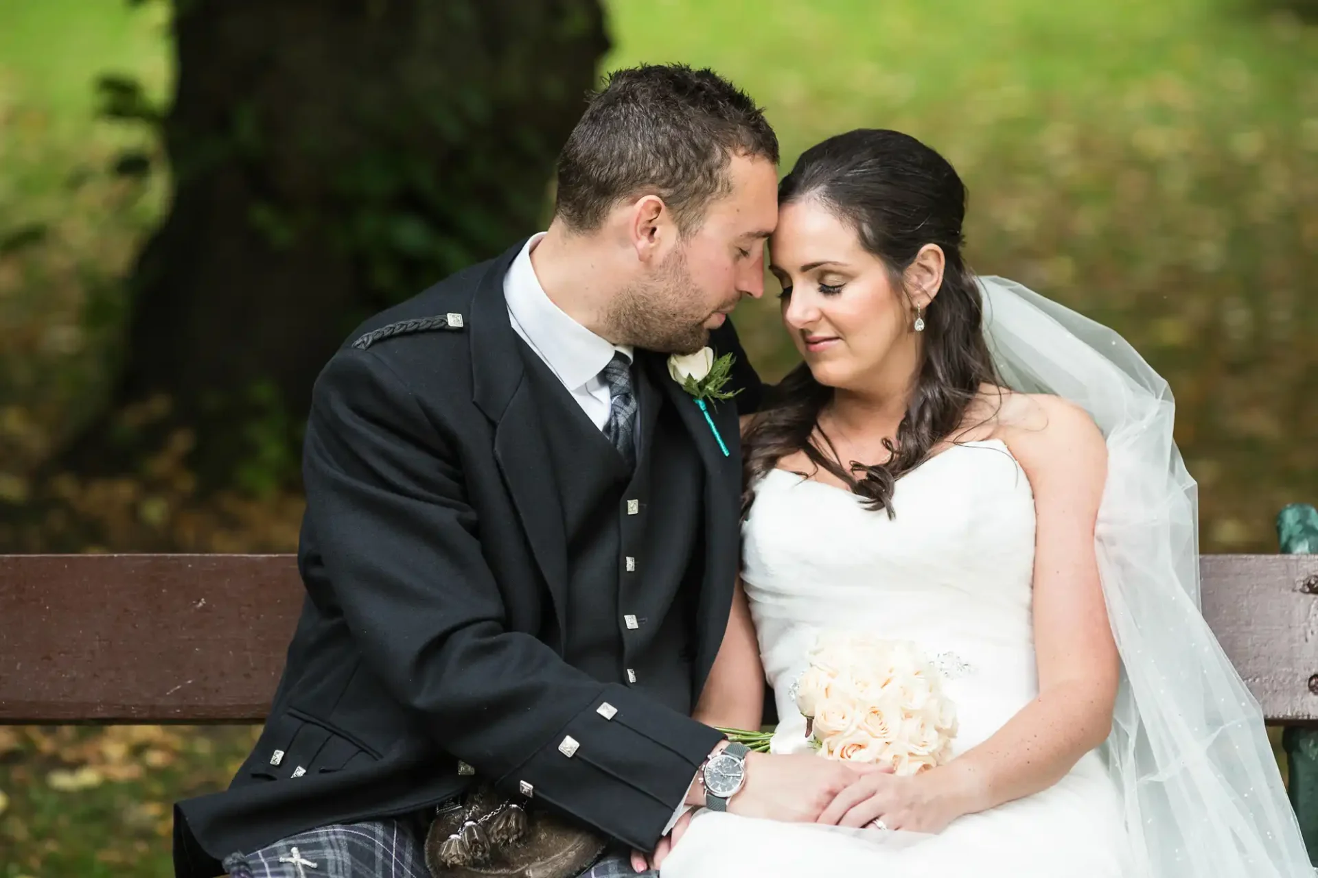 A bride in a white dress and a groom in a kilt share an intimate moment on a park bench, surrounded by greenery.