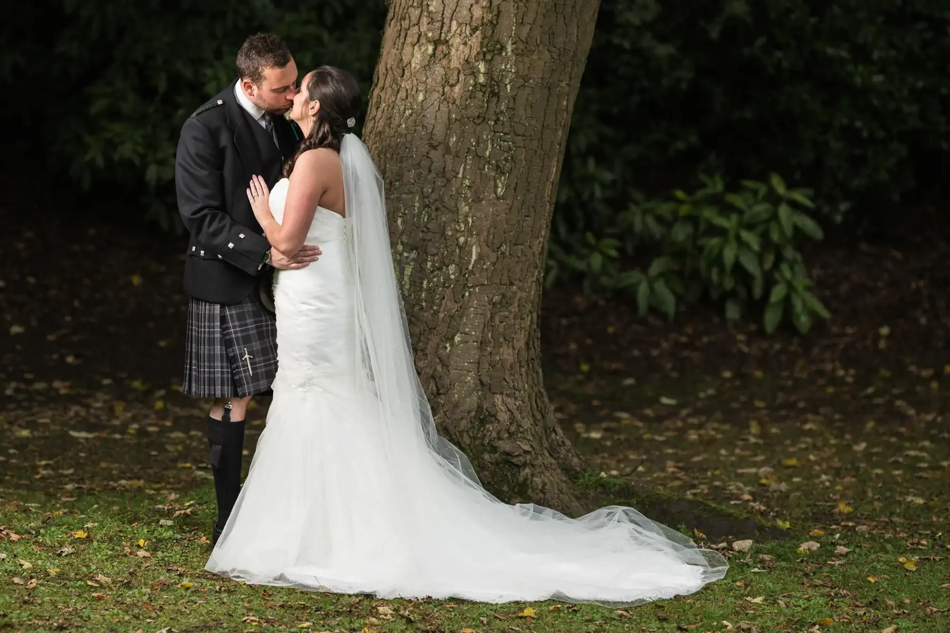 A bride in a long white dress and a groom in a kilt share a kiss under a tree in a park, surrounded by fallen leaves.