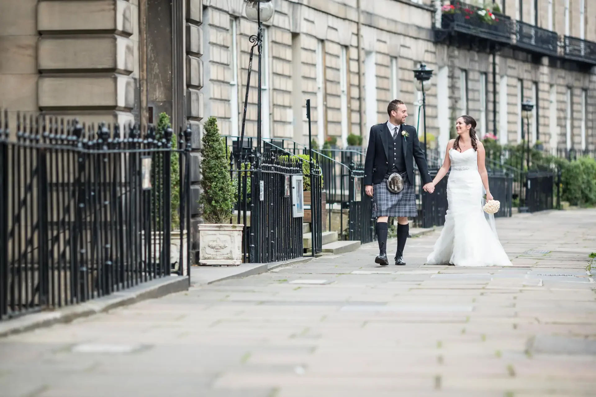 Bride and groom walking hand in hand along a city street, with the groom wearing a kilt and the bride in a white dress.