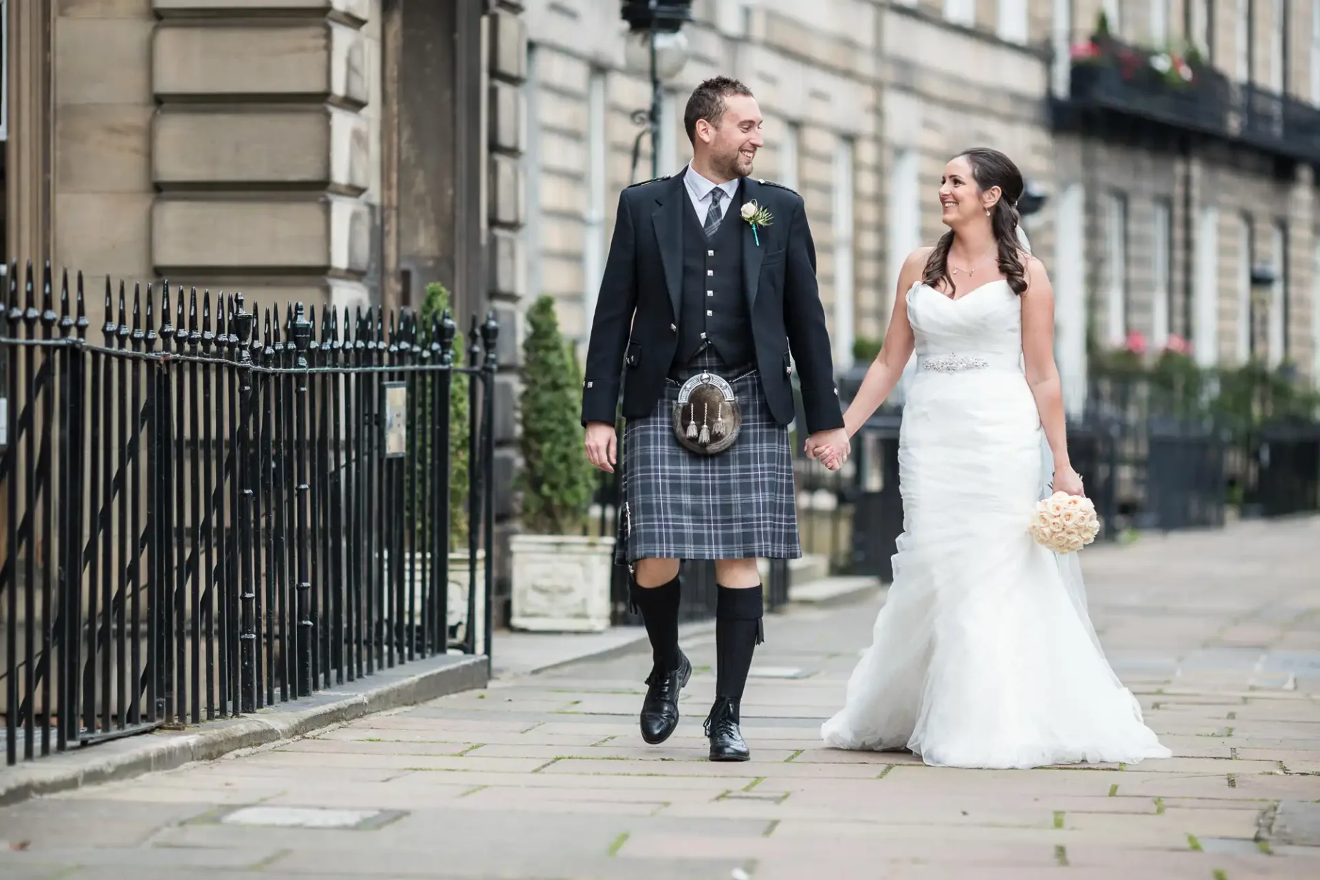 A newlywed couple smiling and walking down a city street, the groom wearing a kilt and the bride in a white gown.