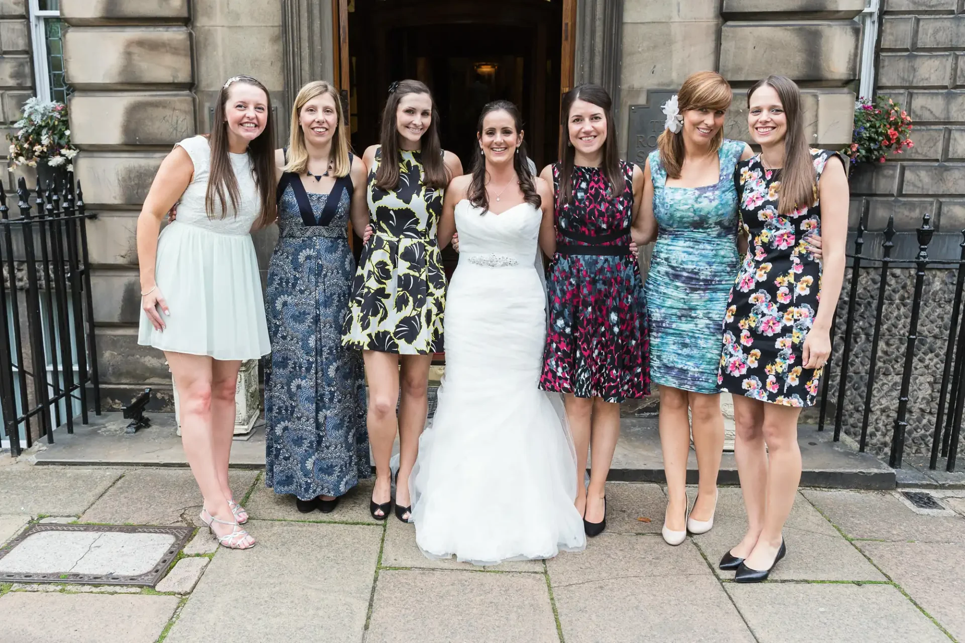 Seven women in dresses, including one in a white bridal gown, smiling and posing together on a cobblestone street.