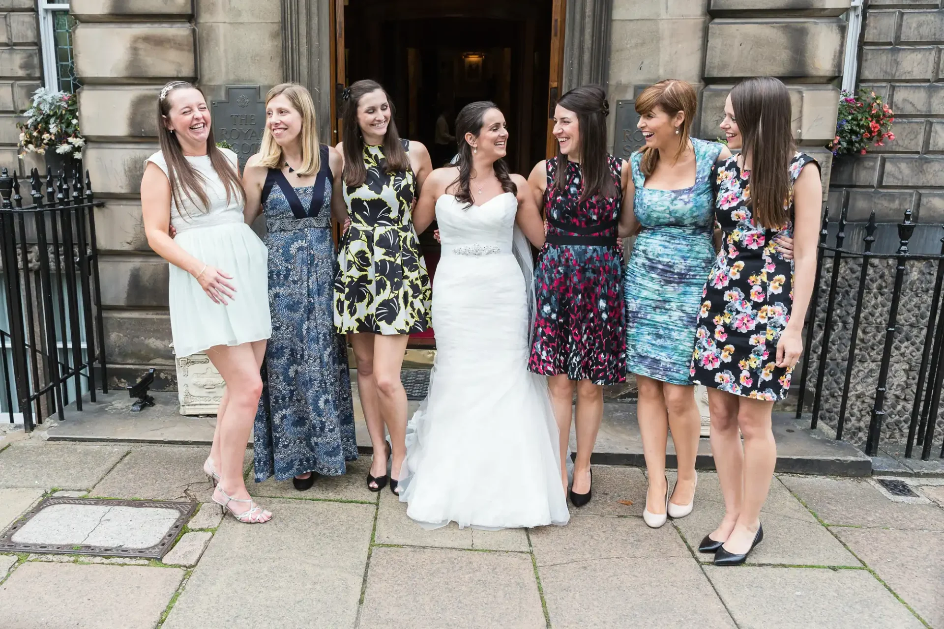A bride in a white dress and six women in various cocktail dresses pose smiling on a stone walkway outside a building.