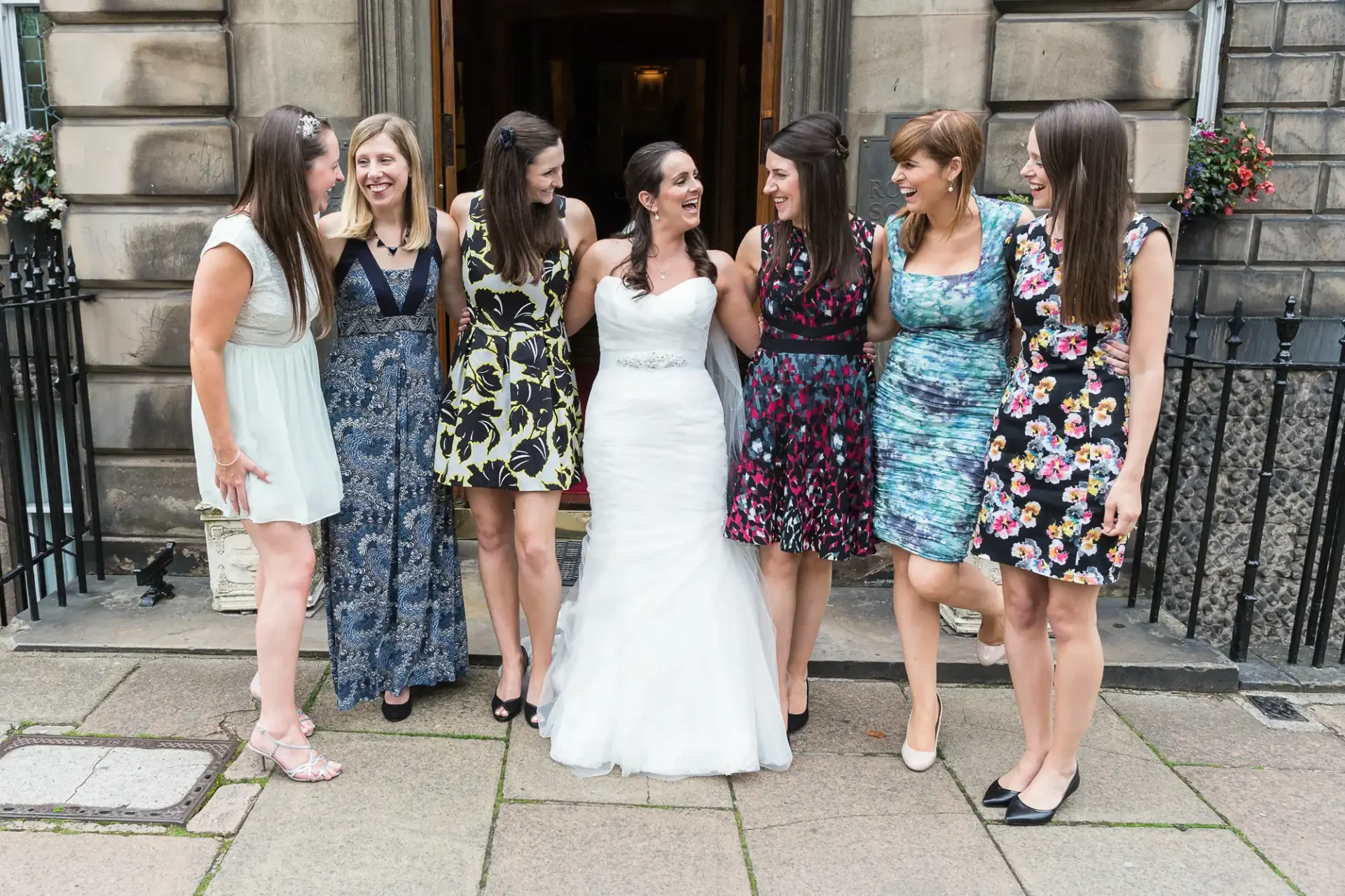 A bride in a white dress laughs with six women in floral and solid-colored dresses outside a building.