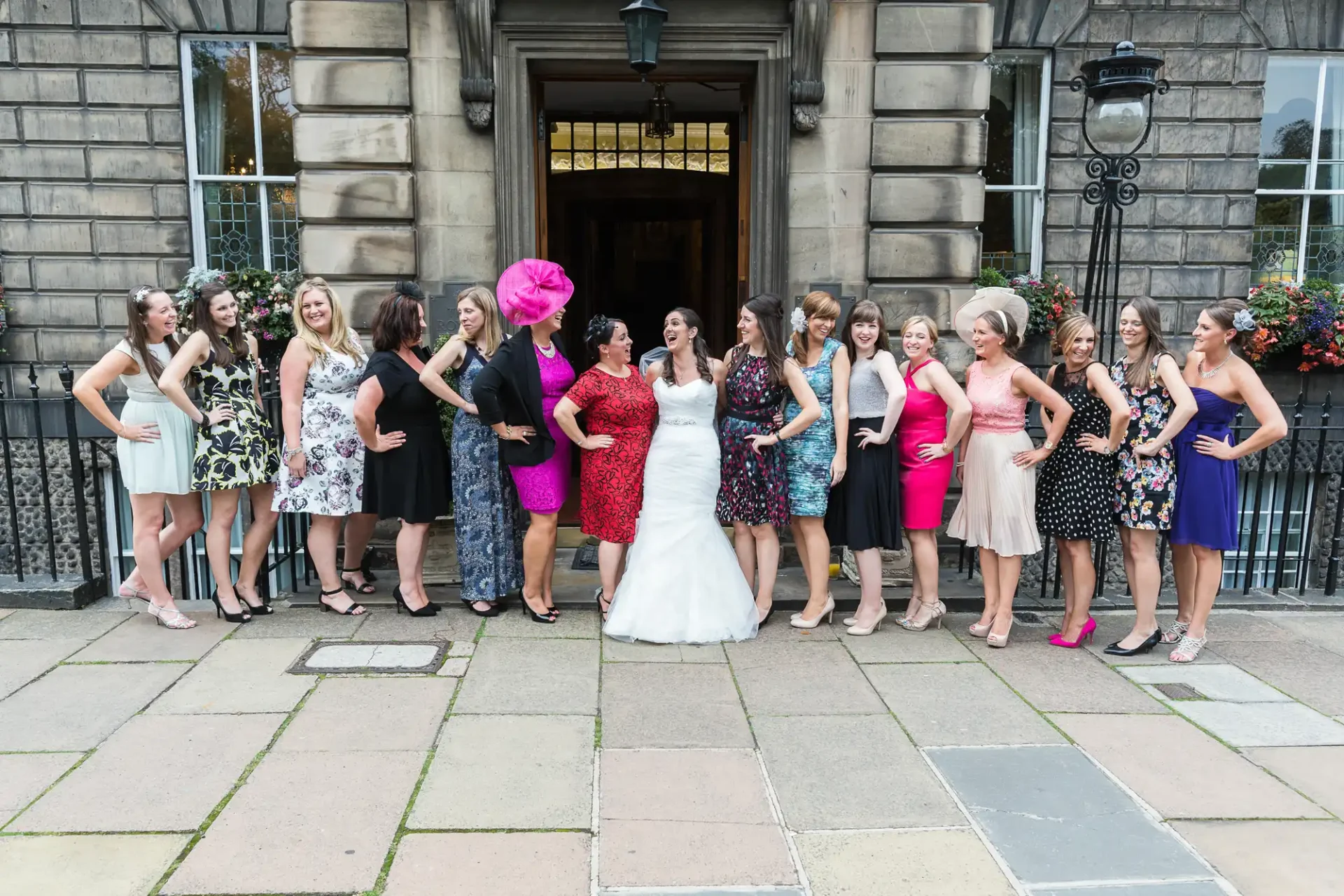 A group of women in formal attire posing happily outside a building, with one in a white wedding dress.