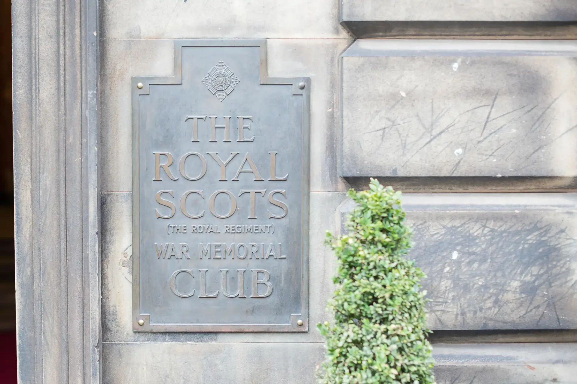 Bronze plaque labeled "the royal scots (the royal regiment) war memorial club" mounted on a stone wall beside a shrub.