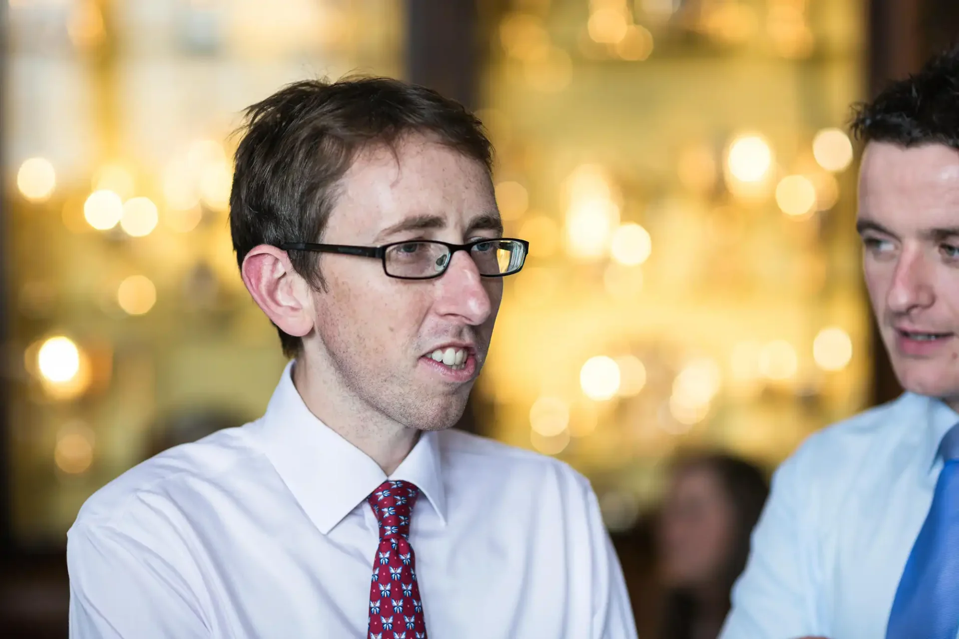 Two men in business attire engaging in conversation, one wearing glasses and speaking animatedly in a restaurant with blurred background.