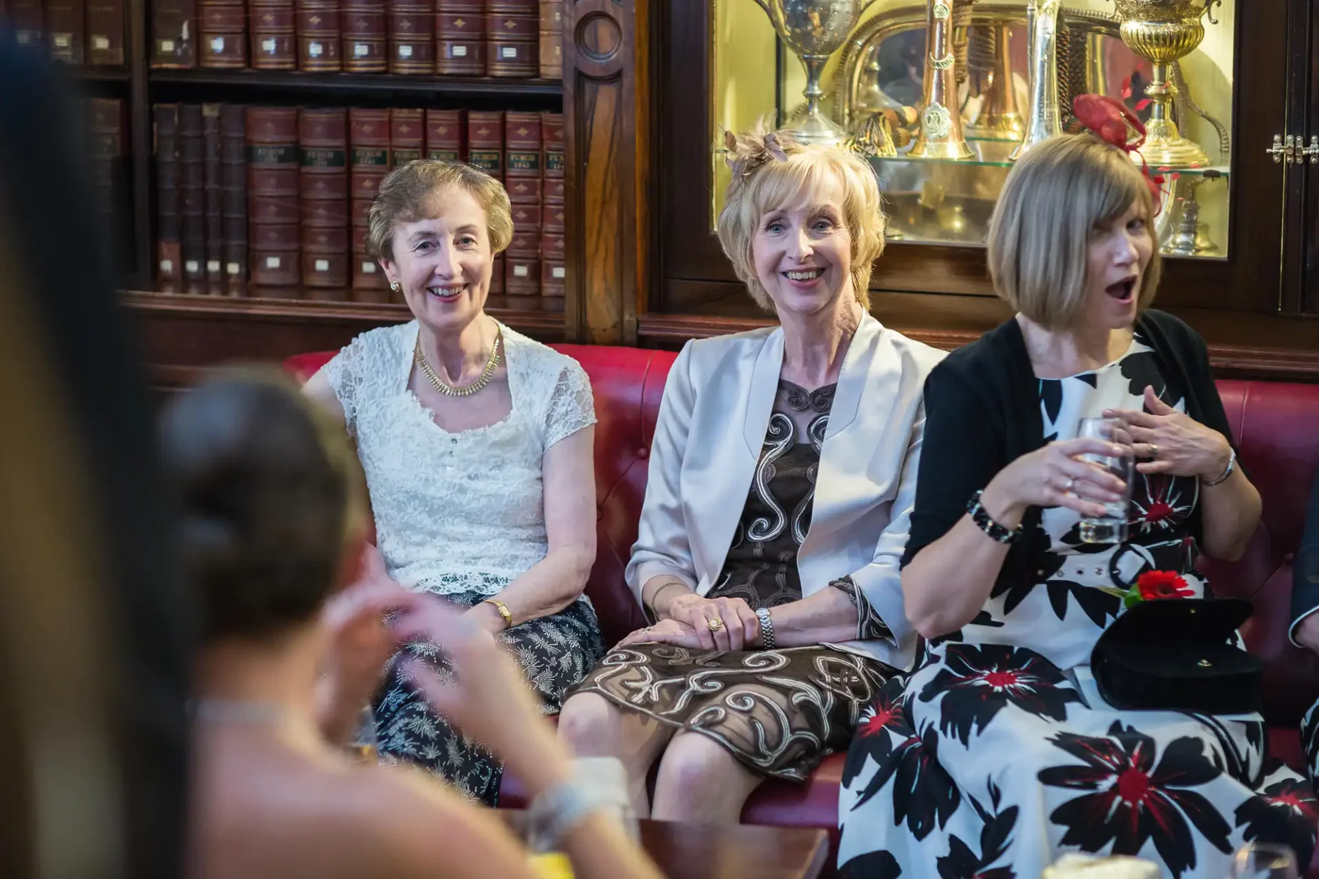 Three older women seated in a library, smiling and engaged in conversation at a social gathering, elegantly dressed.