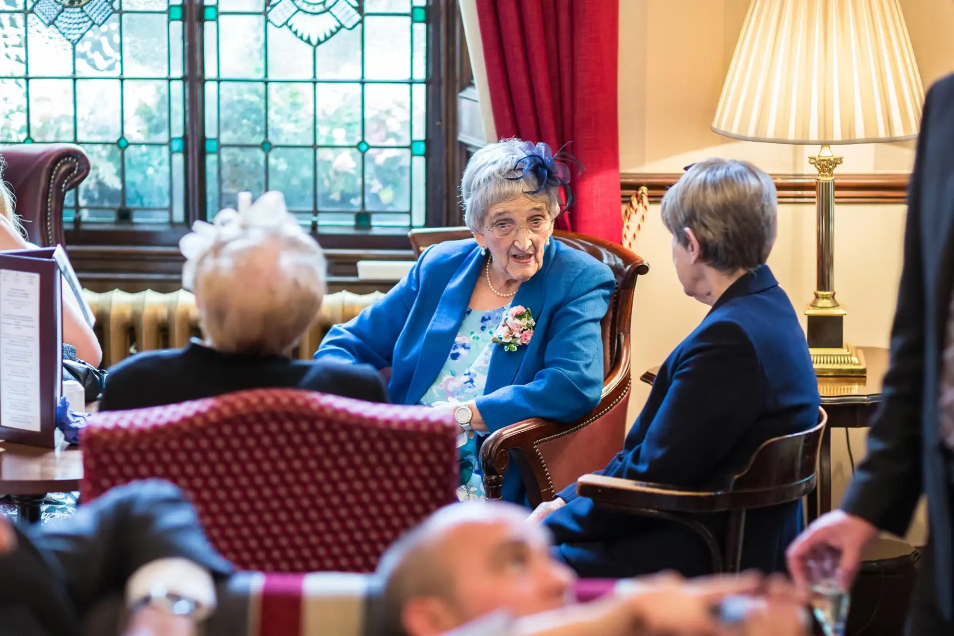 Elderly woman in blue jacket and floral corsage talking to a man in a suit at a social gathering in a room with classic decor.