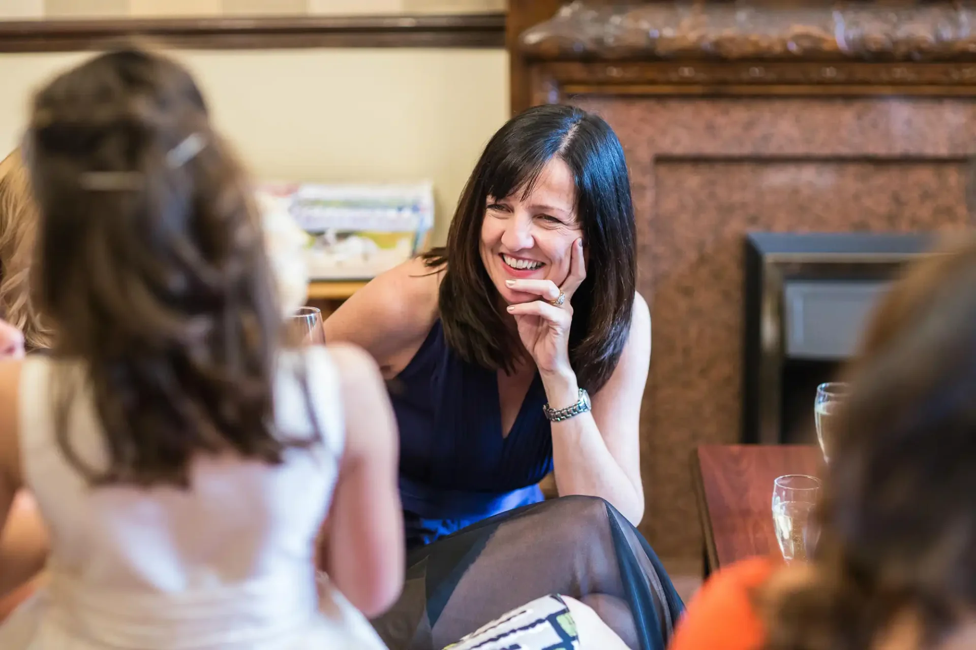 A woman with dark hair laughs joyfully during a conversation with three other women around a table in a warmly lit room.
