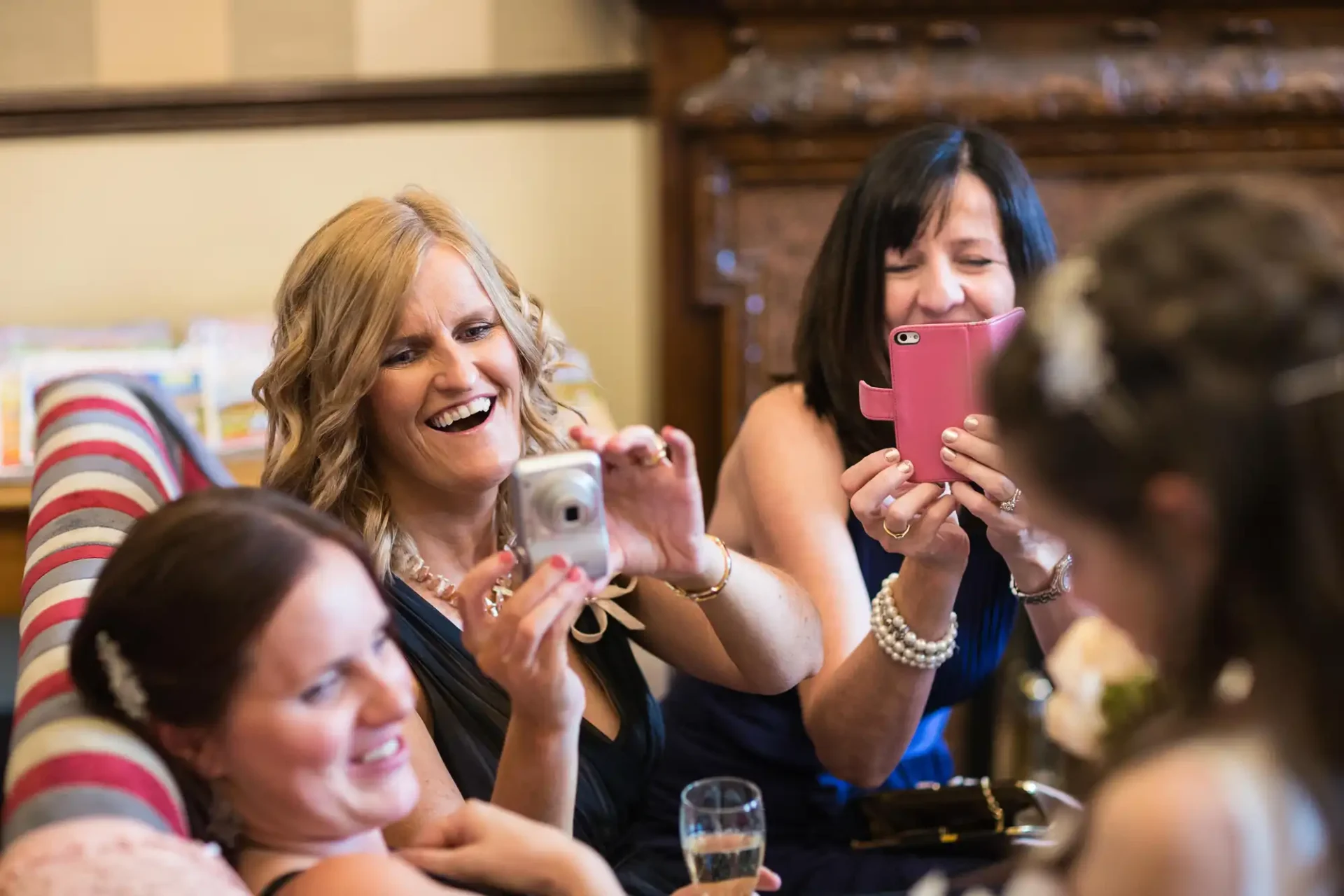 Two women smiling and capturing photos with their phones at a social gathering, surrounded by other guests enjoying the event.