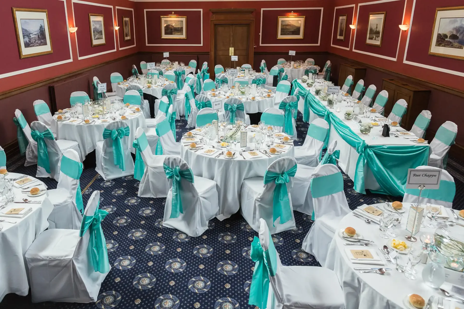 Elegant dining hall set for an event with round tables, white chairs adorned with teal sashes, and formal table settings.