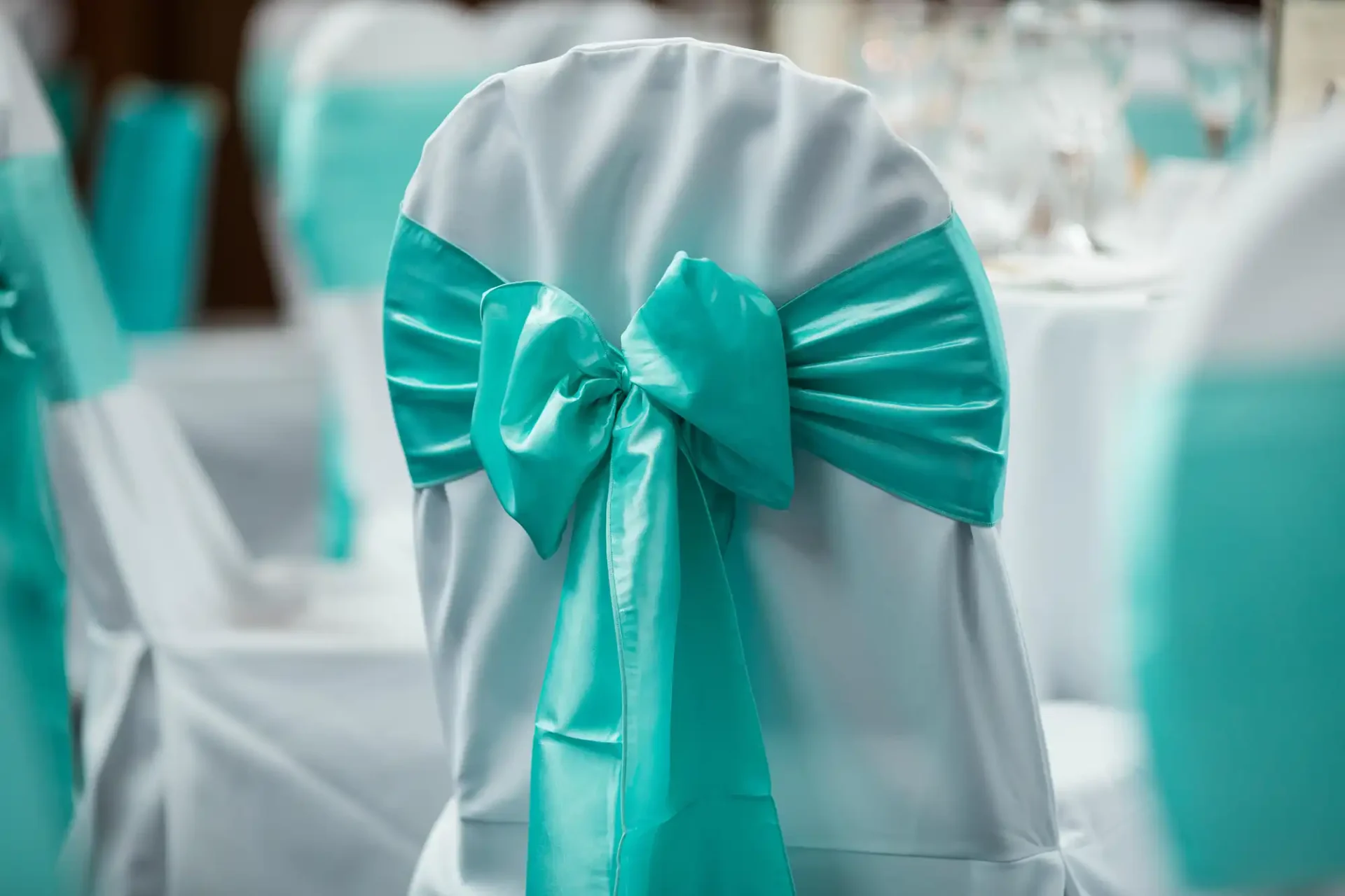 Elegant white chair with a teal satin sash tied in a bow, set up at a decorated event venue with more similarly adorned chairs and tables in the background.