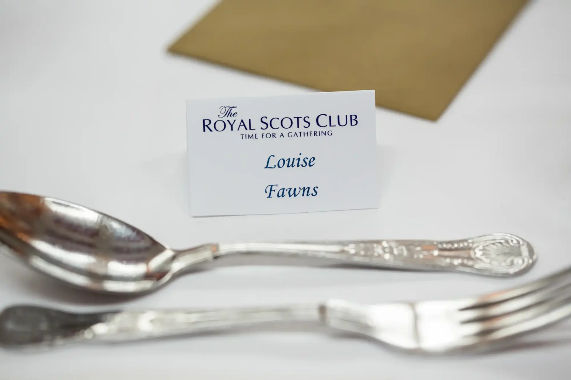 Place card reading "louise fawns" at a dining setting with silverware and a golden envelope on a white tablecloth.