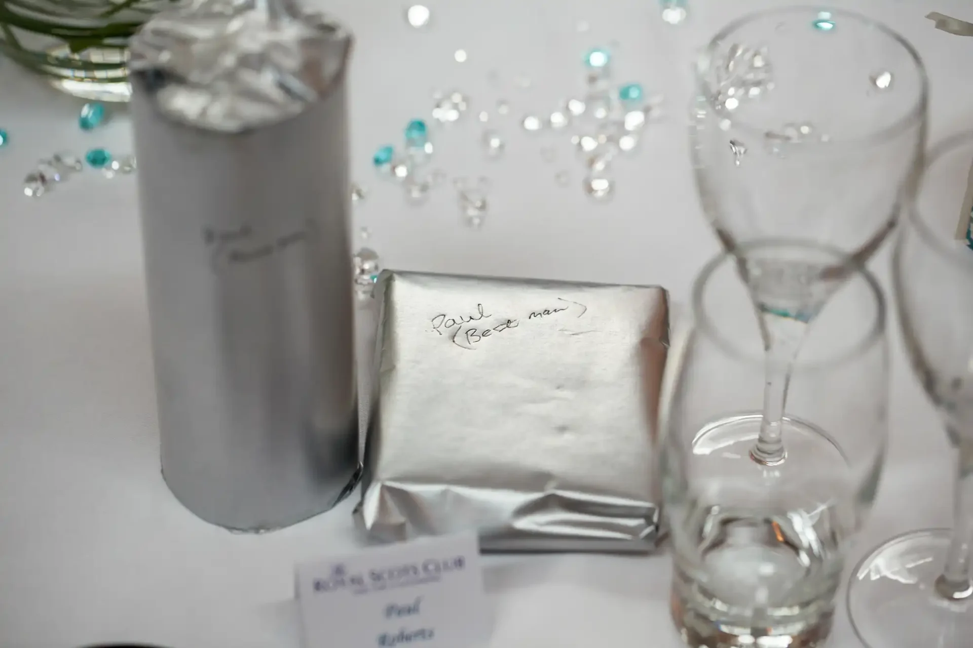 Table setting with a foil-wrapped gift labeled "paul's present" beside scattered decorative crystals and glassware.