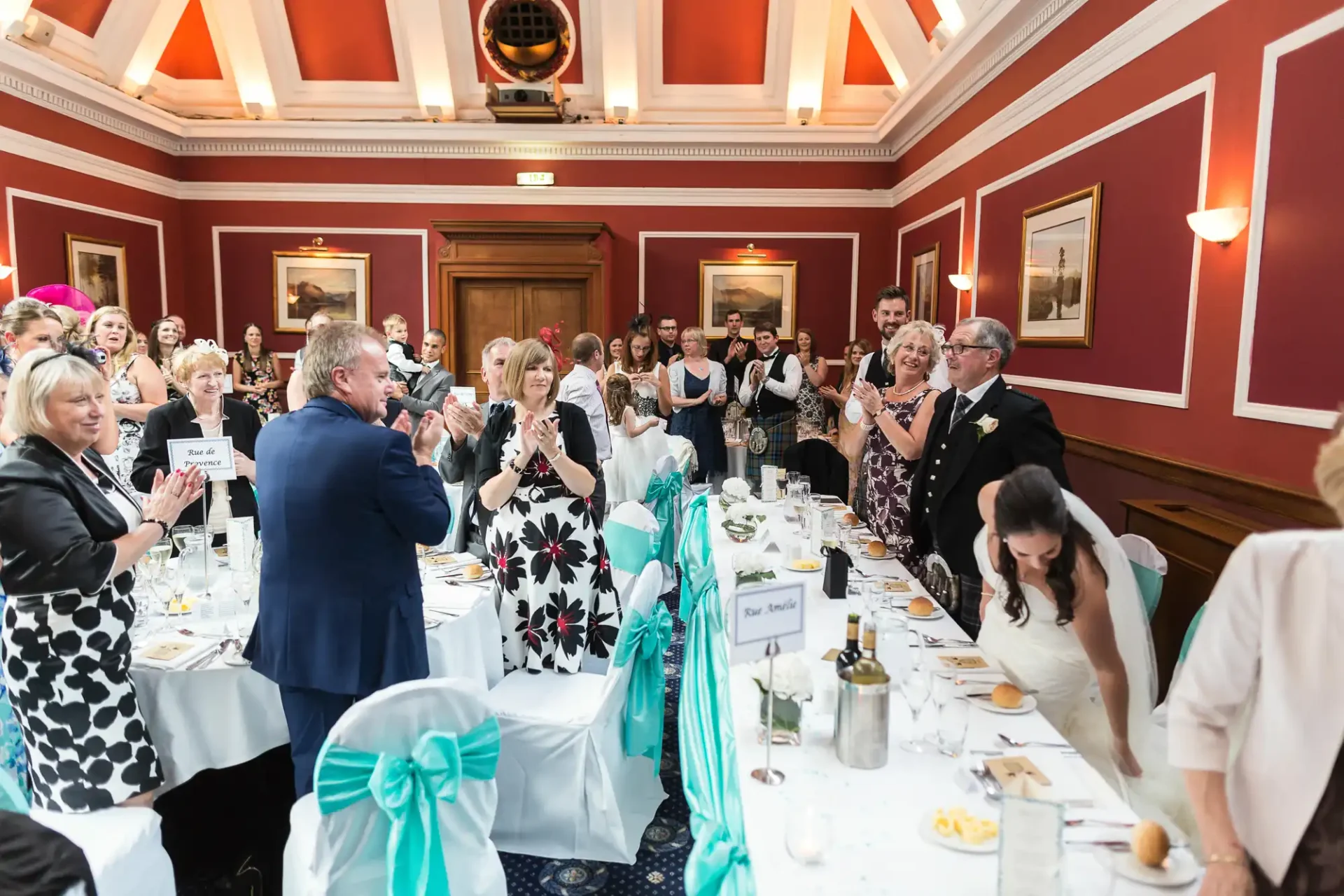 Wedding guests applauding as a couple walks through a dining hall decorated with teal accents.